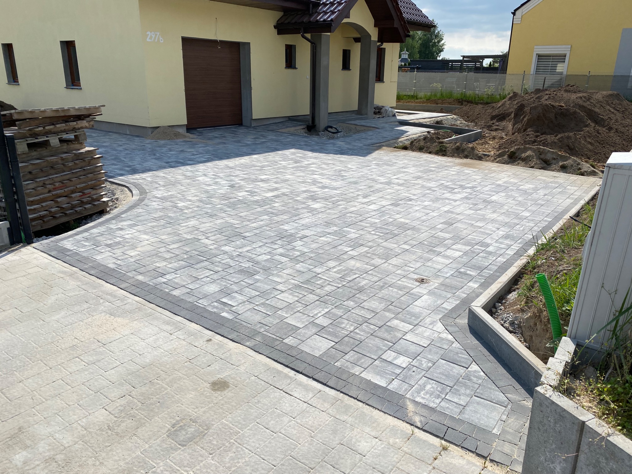 Nowoczesny podjazd z szarej kostki brukowej ułożonej we wzór jodełki, z obrzeżem z ciemniejszych kostek, przed domem we Wrocławiu. Widoczne palety i przygotowane podłoże pod dalsze prace.