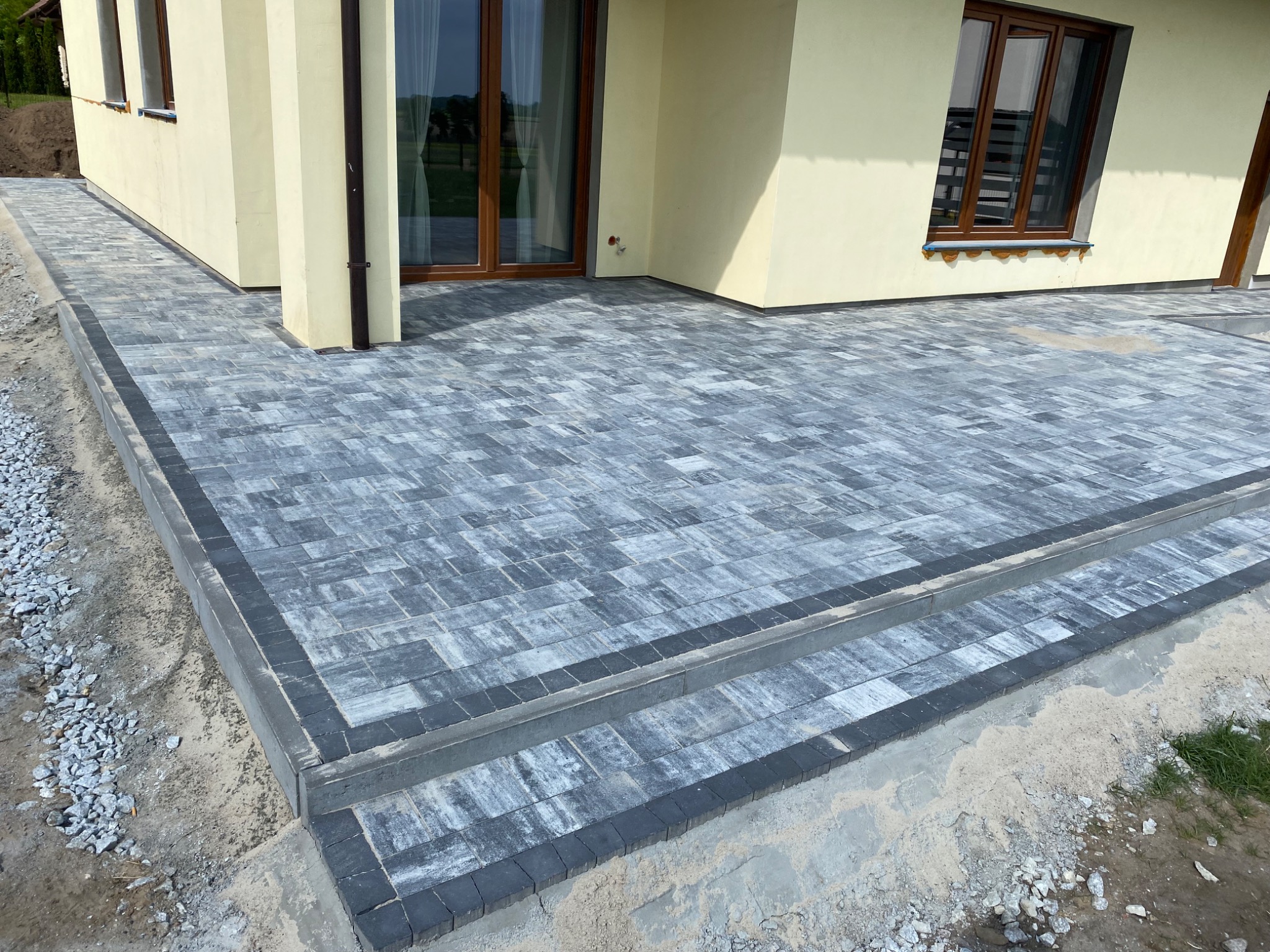 Nowoczesny taras z szarej kostki brukowej ułożonej wzorem prostokątnym, z ciemnoszarą obwódką, prowadzący do domu z beżową elewacją i brązowymi oknami.