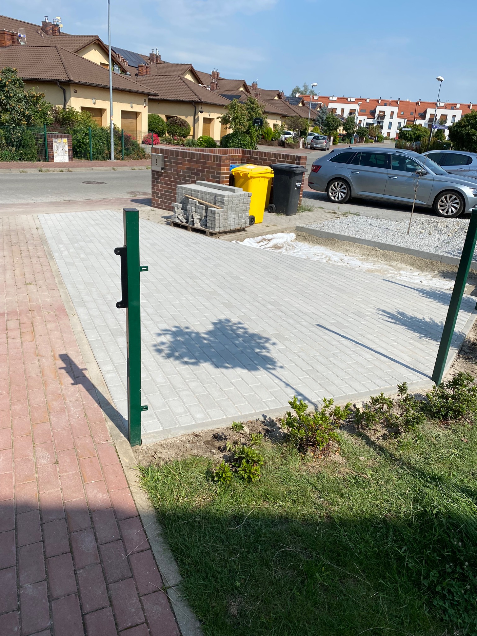 Nowo ułożona szara kostka brukowa na podjeździe przed domem, widoczne palety z kostką, obrzeża i słupki ogrodzeniowe.