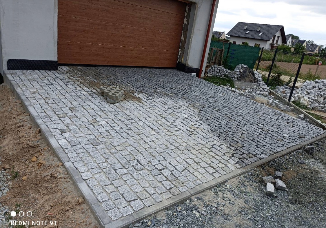 Nierównomiernie ułożona szara kostka granitowa przed garażem, widoczne pozostałości piasku i gruzu, w tle domy jednorodzinne.