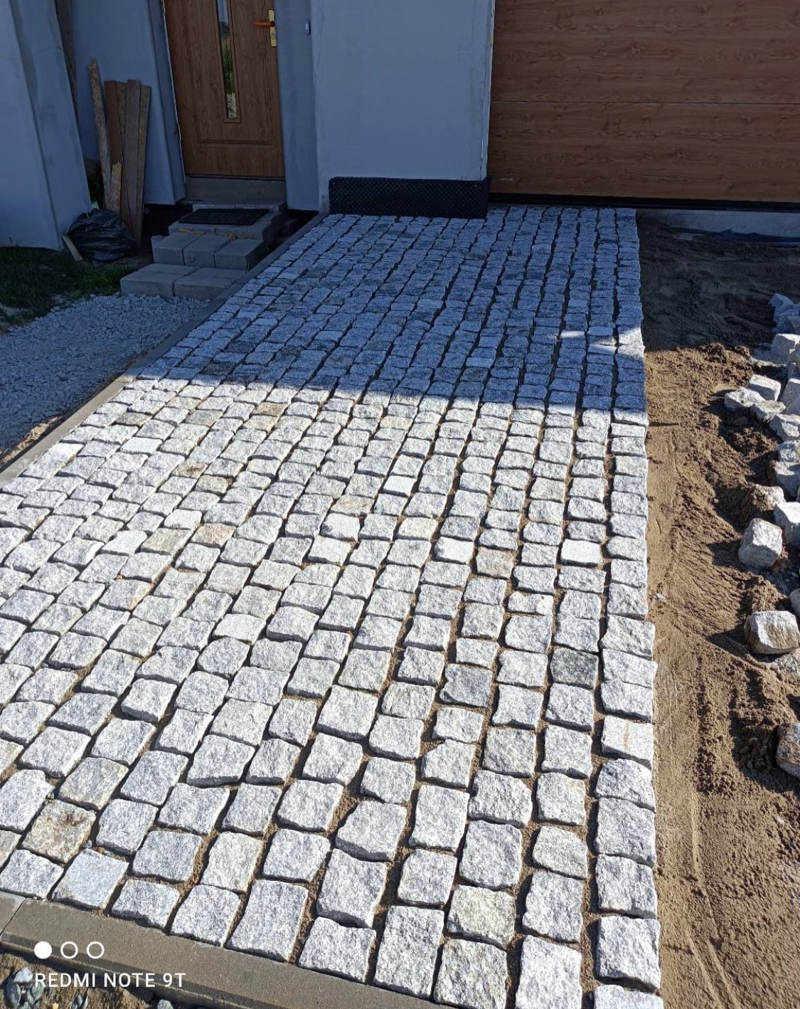 Świeżo ułożony chodnik z jasnoszarej kostki granitowej prowadzący do drzwi wejściowych domu, z widocznymi pozostałościami piasku na łączeniach i przygotowanym obszarem do dalszego układania.
