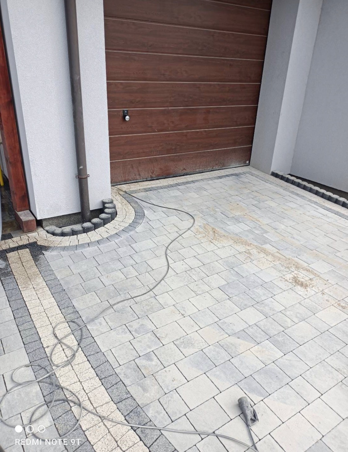 Ułożona kostka brukowa w odcieniach szarości i beżu przed garażem z brązową bramą, widoczny przewód elektryczny i rynna.