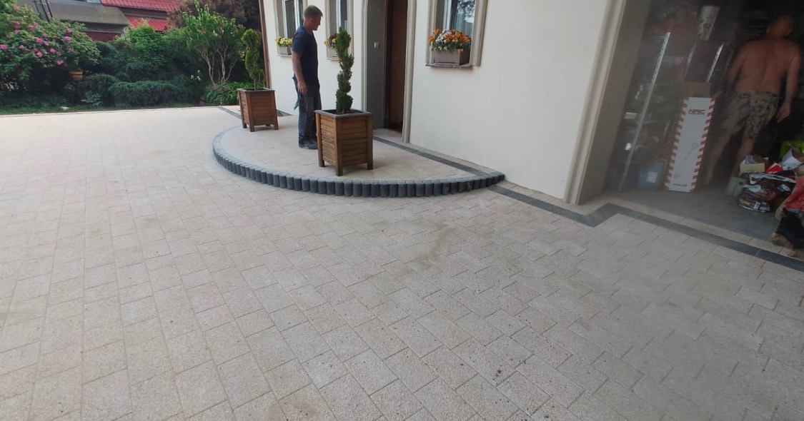 Jasna kostka brukowa ułożona na podjeździe przed domem z obrzeżem z ciemniejszych kostek, donice z drzewkami, widoczna osoba w tle.