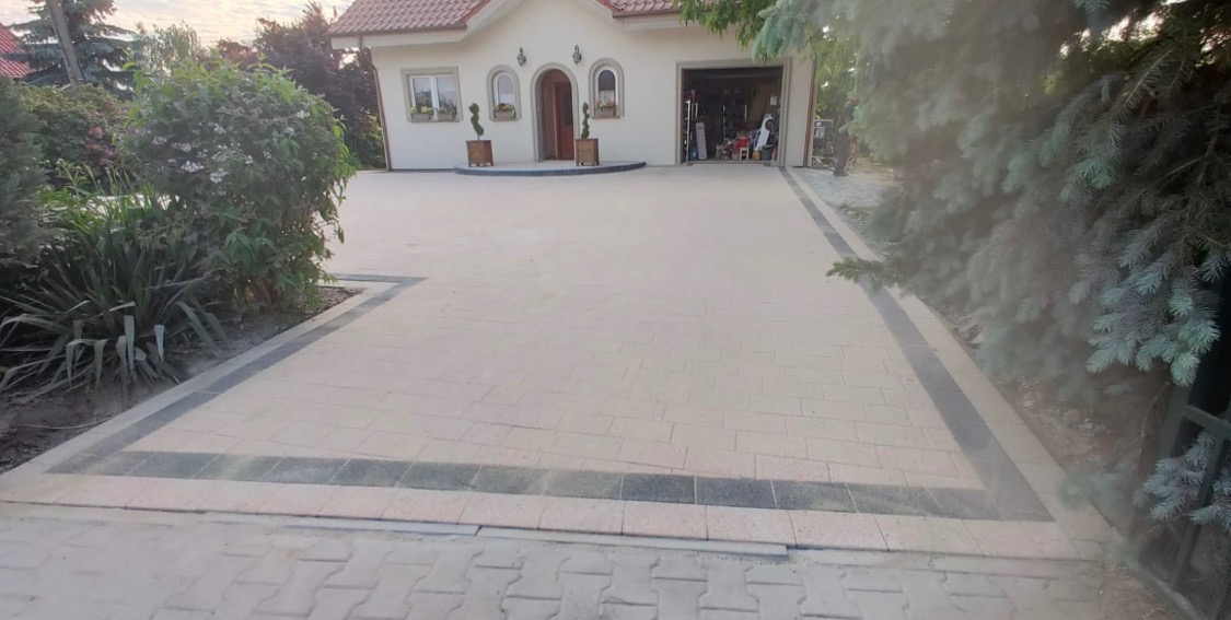 Jasna kostka brukowa ułożona na podjeździe do domu jednorodzinnego z garażem, obrzeża z ciemnej kostki, widok z poziomu gruntu.