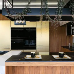 Nowoczesna kuchnia z drewnianymi i złotymi szafkami, czarnym blatem i wiszącymi roślinami. Na blacie stoją dwie filiżanki na dekoracyjnych talerzykach.