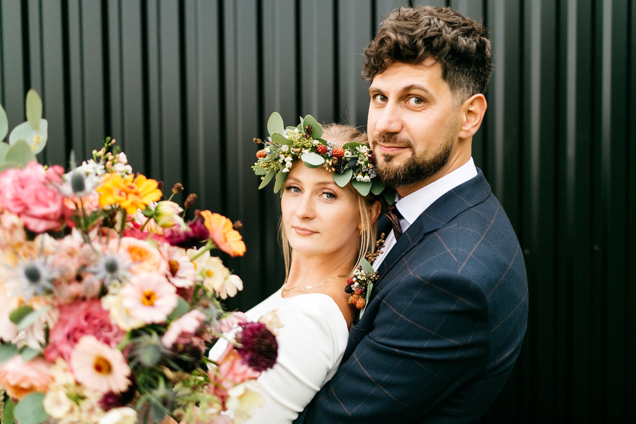Para młoda w objęciach na tle ciemnej, pionowo żebrowanej ściany; Panna młoda w wianku z kwiatów i jagód, trzyma bukiet różnokolorowych kwiatów.