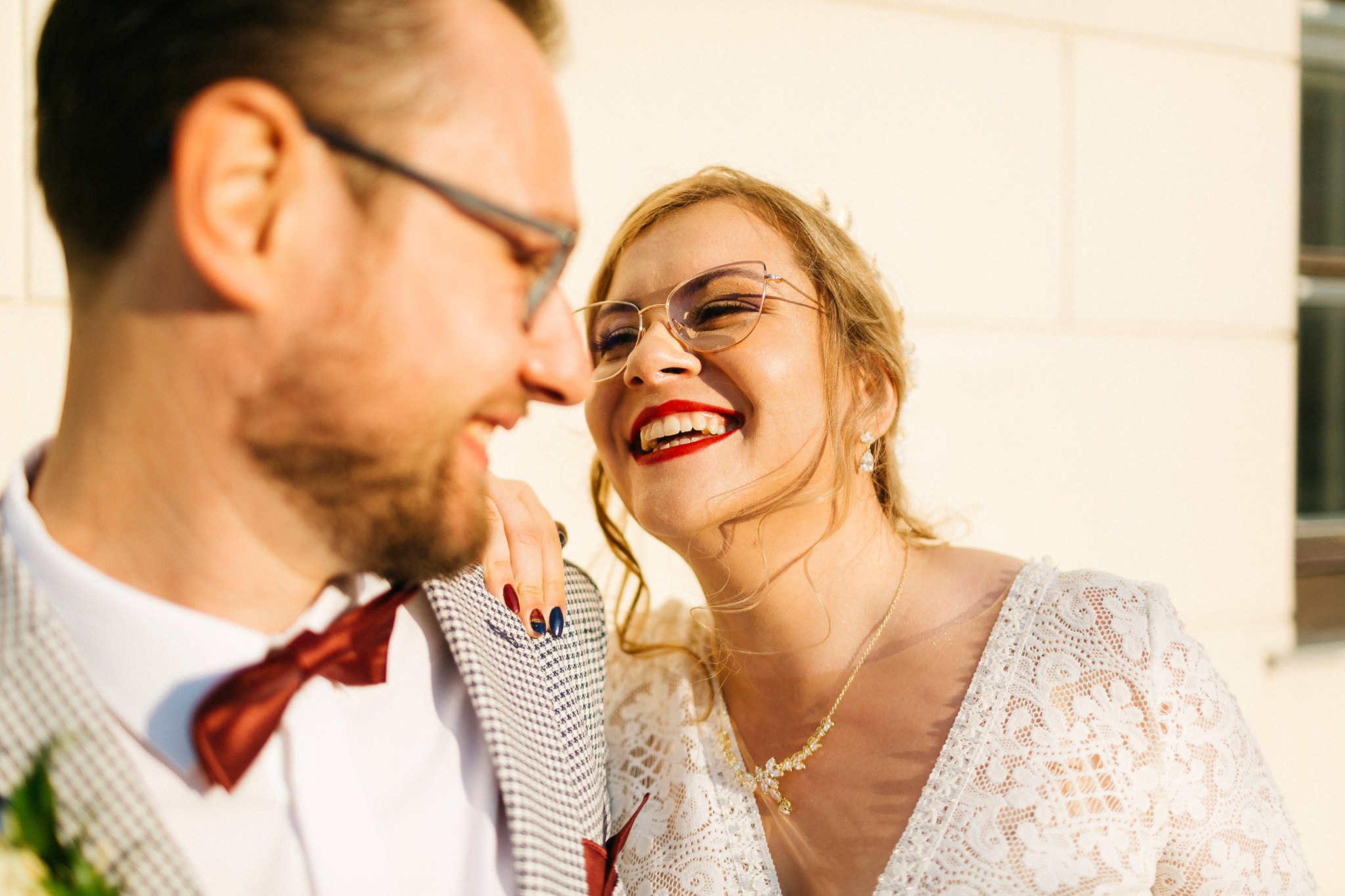 Uśmiechnięta para młoda w okularach, panna młoda z czerwonymi ustami i manicure w kolorach granatu i bordo, opiera rękę na ramieniu pana młodego w muszce.