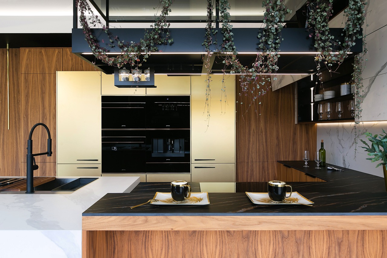 Nowoczesna kuchnia z drewnianymi i złotymi szafkami, czarnym blatem i wiszącymi roślinami. Na blacie stoją dwie filiżanki na dekoracyjnych talerzykach.