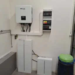 Biała instalacja fotowoltaiczna SOLAX z magazynem energii, zamontowana na ścianie w pomieszczeniu. Widoczny inwerter, moduły bateryjne, skrzynka z bezpiecznikami i okablowanie.