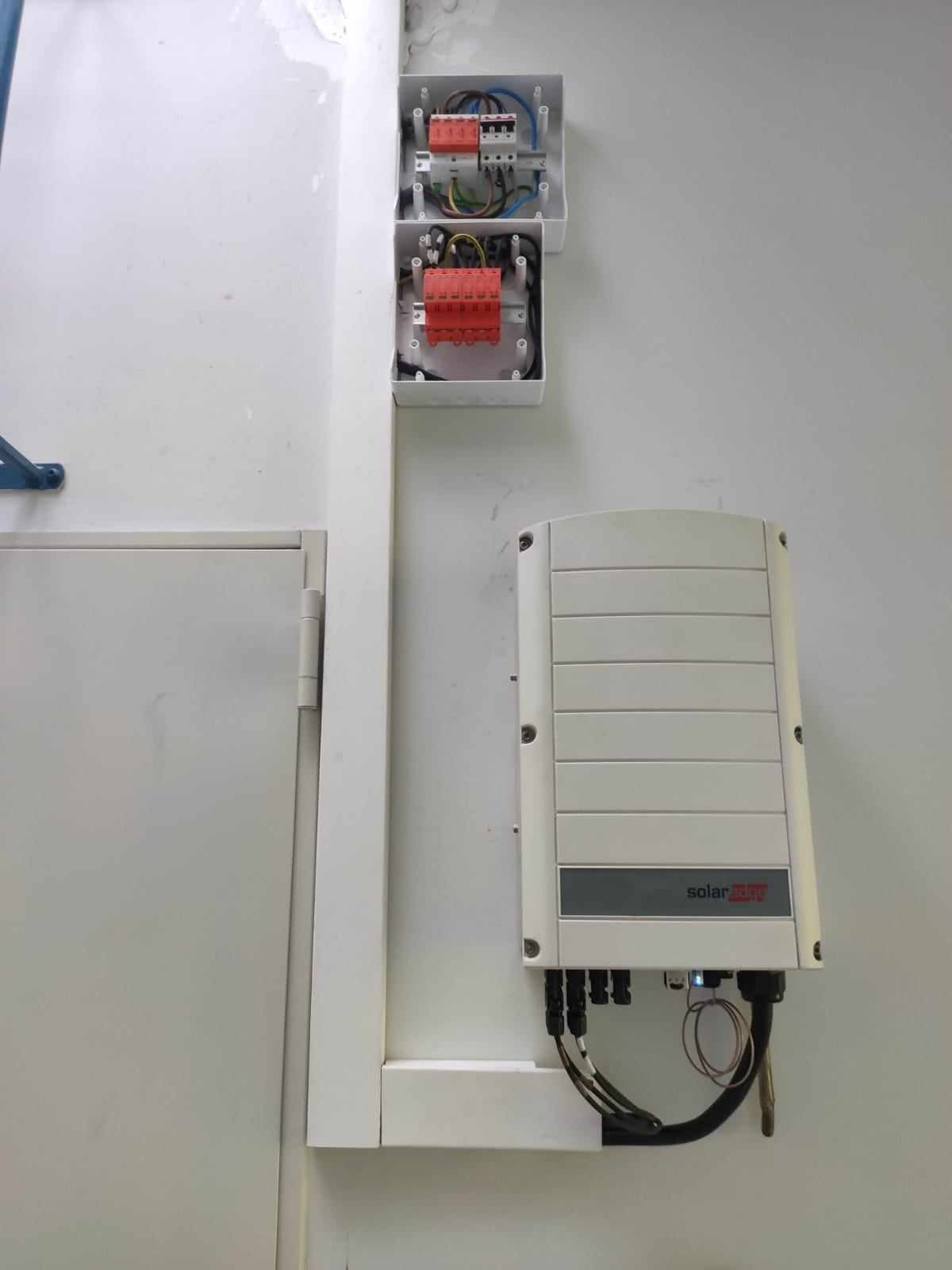 Biała ściana z zamontowaną skrzynką elektryczną z widocznymi bezpiecznikami i przewodami oraz falownikiem SolarEdge z podłączonymi kablami.