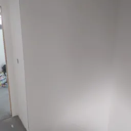 Ściana po nałożeniu jasnej, gładkiej warstwy wykończeniowej, widoczne schody w surowym stanie i fragment torby w tle.