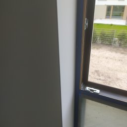 RW remonty i wykończenia wnętrz - Fragment wykończonego wnętrza: gładka, biała ściana sąsiaduje z oknem z niebieską ramą, widok na zewnątrz z zielenią i budynkiem w tle.