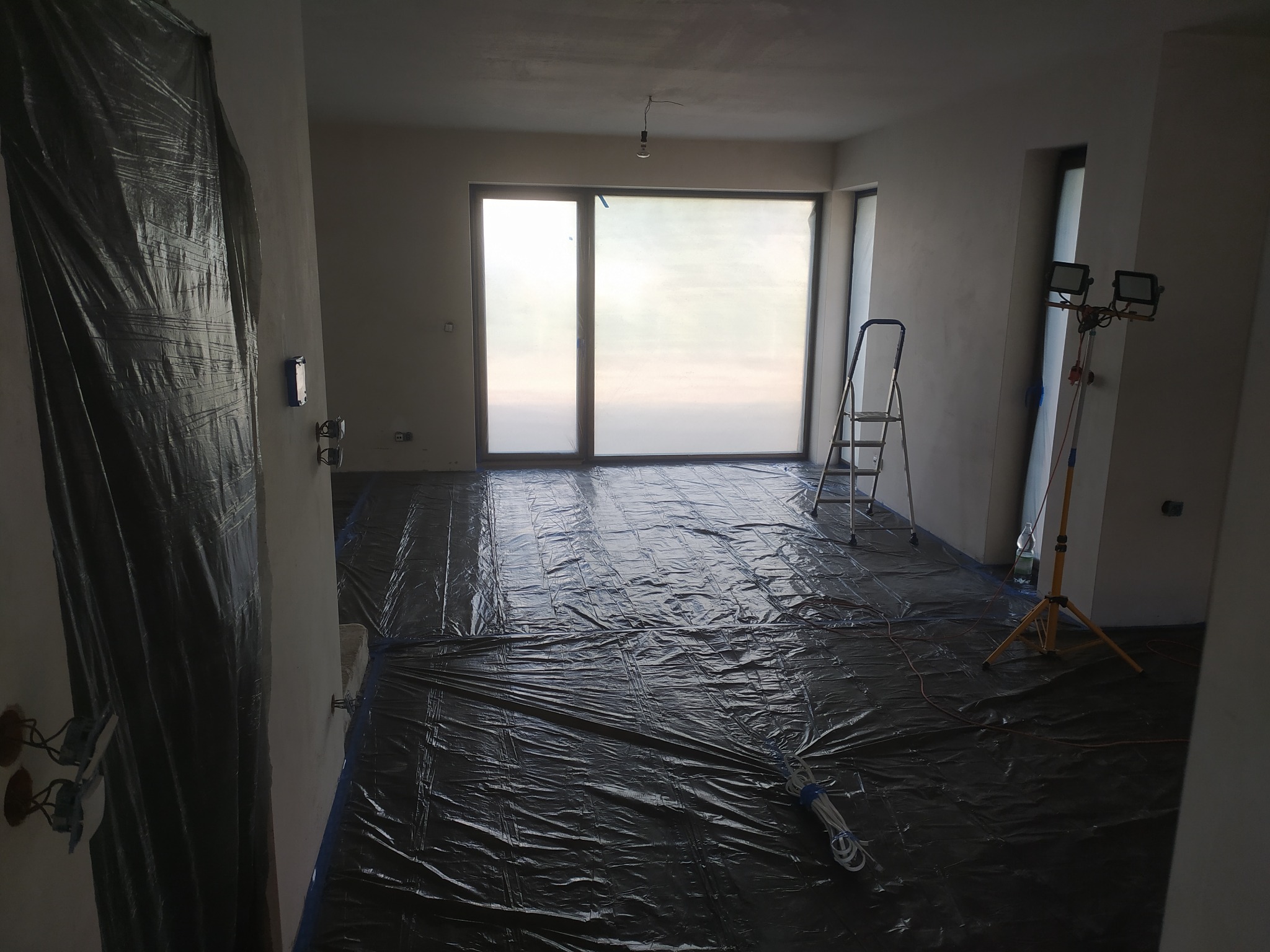 Przygotowane pomieszczenie do prac wykończeniowych: podłoga zabezpieczona folią, ściany wstępnie przygotowane, widoczne narzędzia i oświetlenie budowlane.