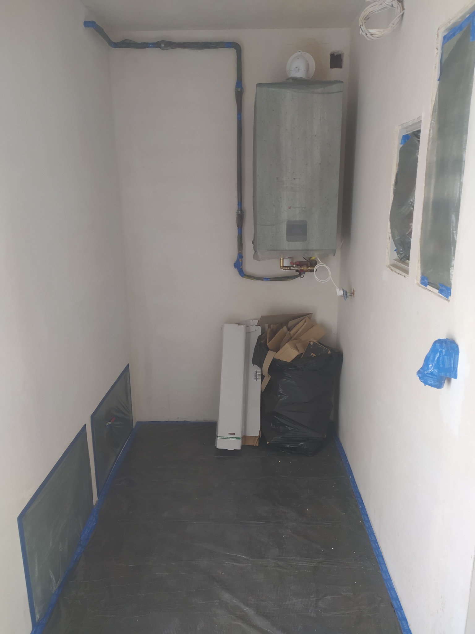 Wnętrze pomieszczenia w trakcie prac remontowych: zabezpieczona podłoga, ściany po nałożeniu warstwy wykończeniowej, widoczny piec gazowy i materiały budowlane.