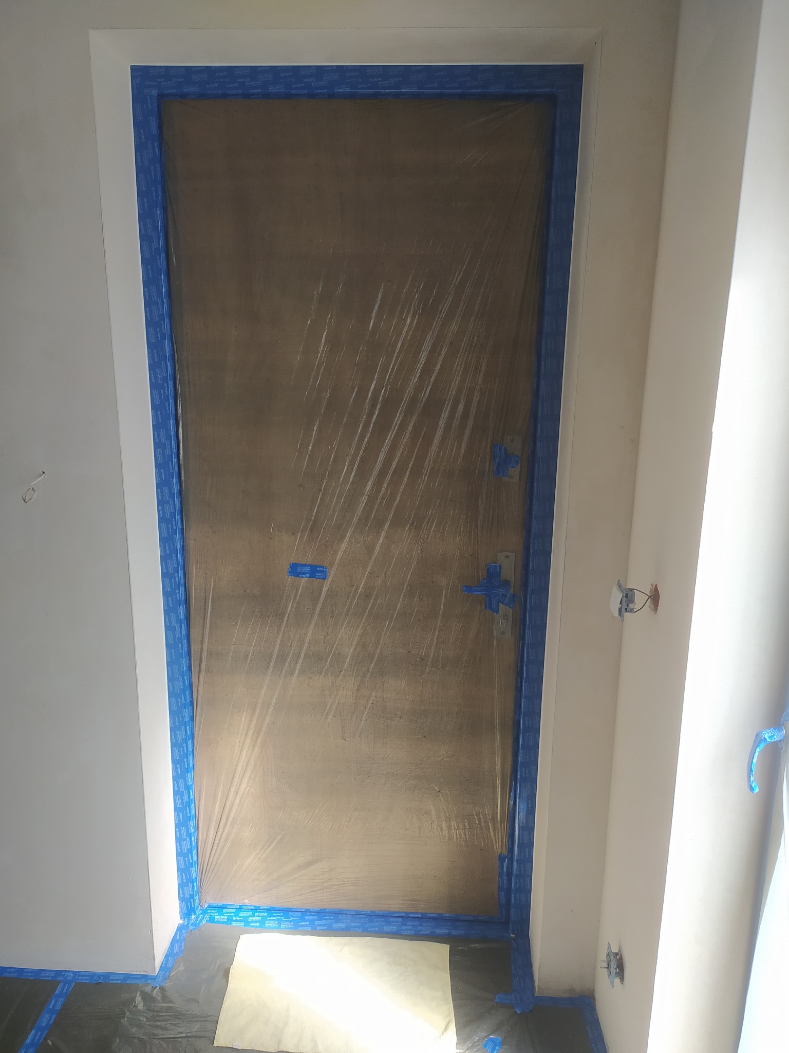 Przygotowanie do malowania: drzwi zabezpieczone folią i niebieską taśmą malarską, ściany częściowo zagruntowane, podłoga osłonięta folią. Widoczny etap prac remontowych.