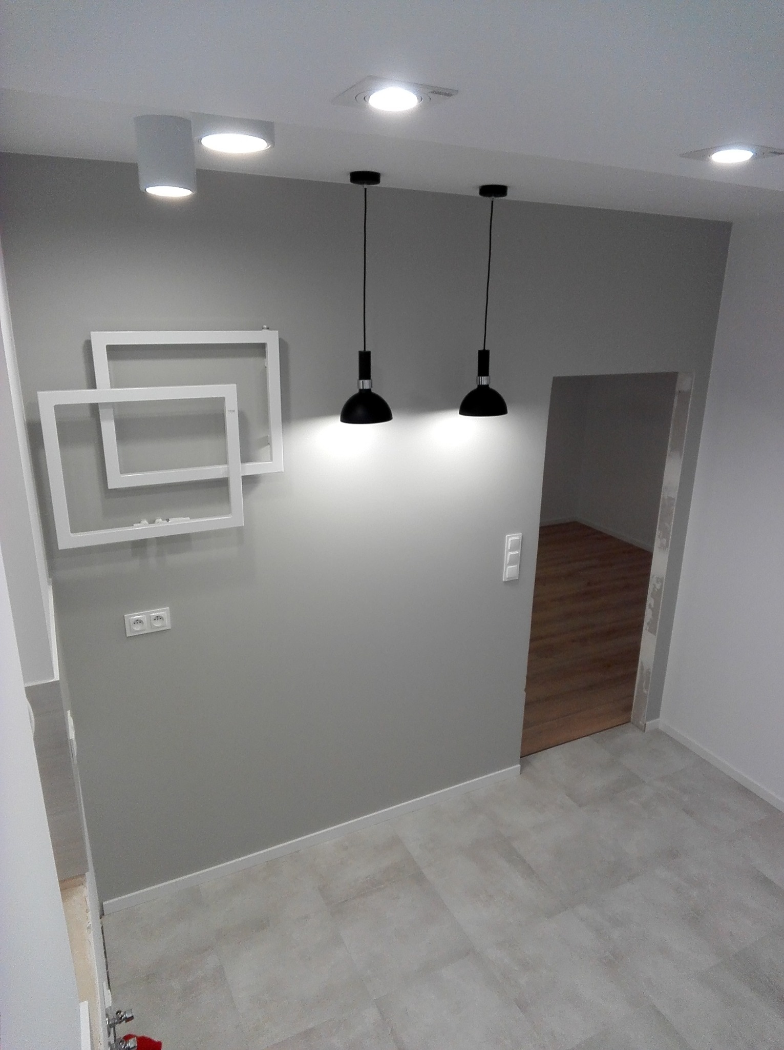 Szara ściana z dekoracyjnymi białymi ramkami, dwiema lampami wiszącymi i gniazdkiem elektrycznym. Widoczny fragment podłogi i wejście do innego pomieszczenia.