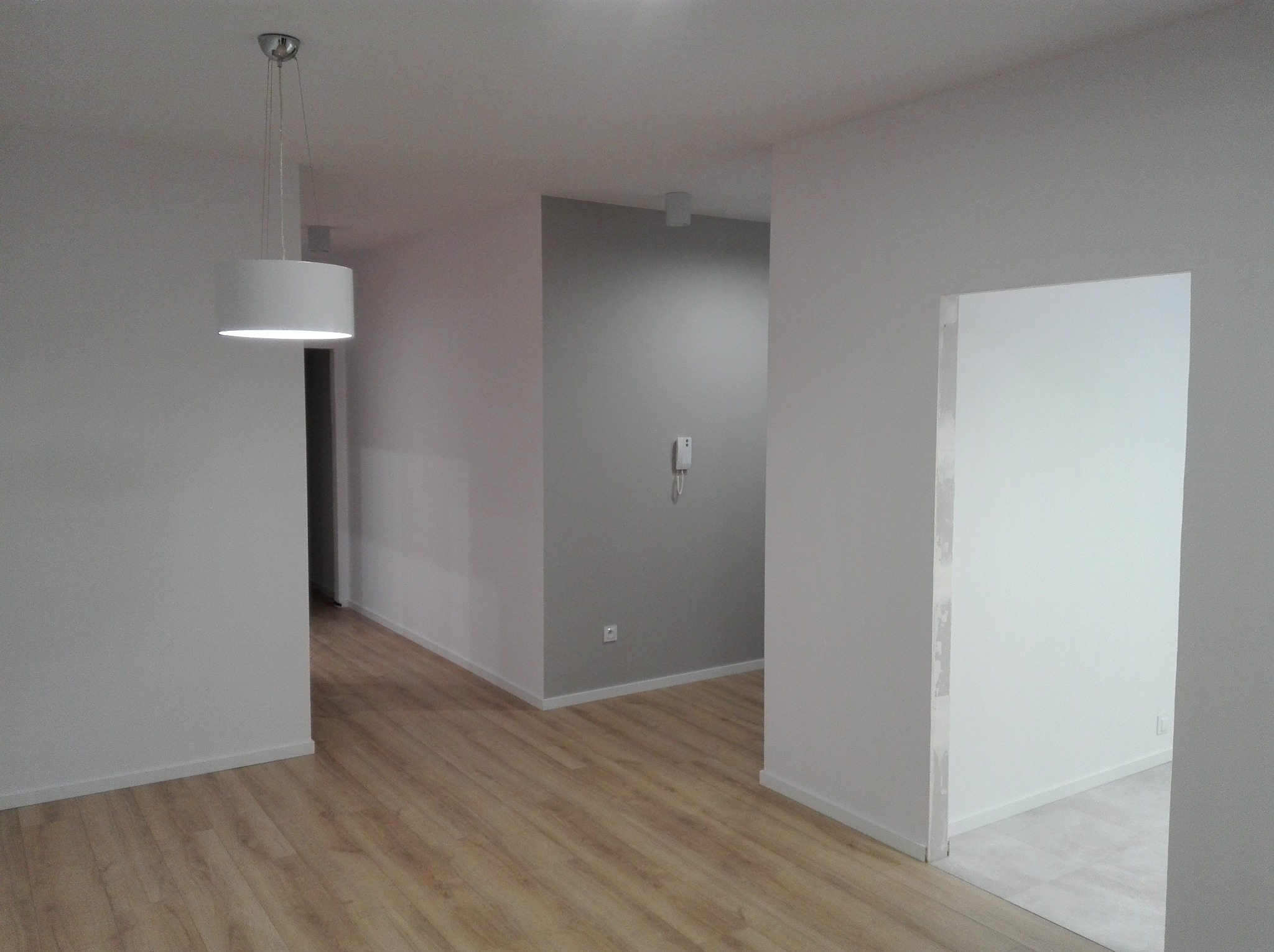 Wnętrze po remoncie: gładkie, białe ściany, drewniana podłoga i nowoczesna lampa wisząca. Fragment ściany w odcieniach szarości. Widoczne gniazdko i domofon.
