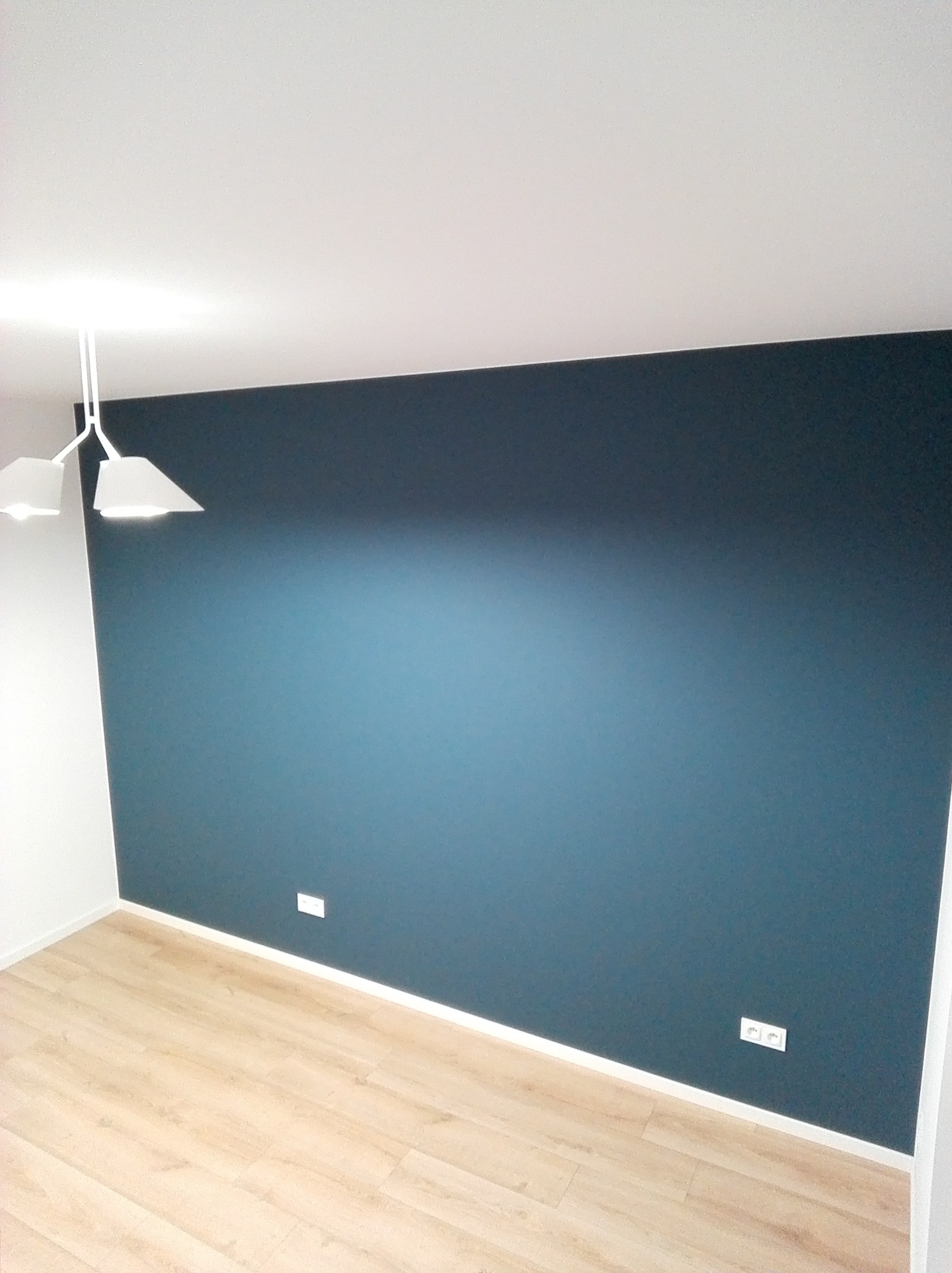 Ściana pomalowana na ciemny odcień niebieskiego, gładka powierzchnia, widoczne gniazdka elektryczne, podłoga z paneli w jasnym odcieniu drewna i białe listwy przypodłogowe.
