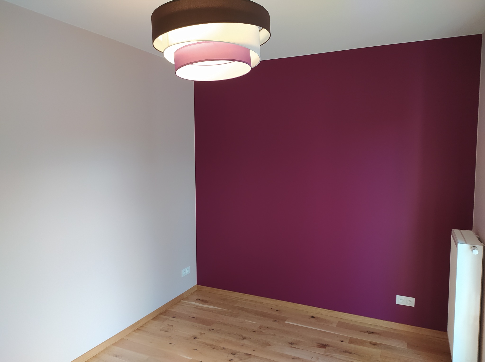 Ściana pomalowana na dwa kolory w pokoju w Warszawie. Jedna ściana biała, druga w odcieniach fioletu. Na suficie lampa. Podłoga drewniana.