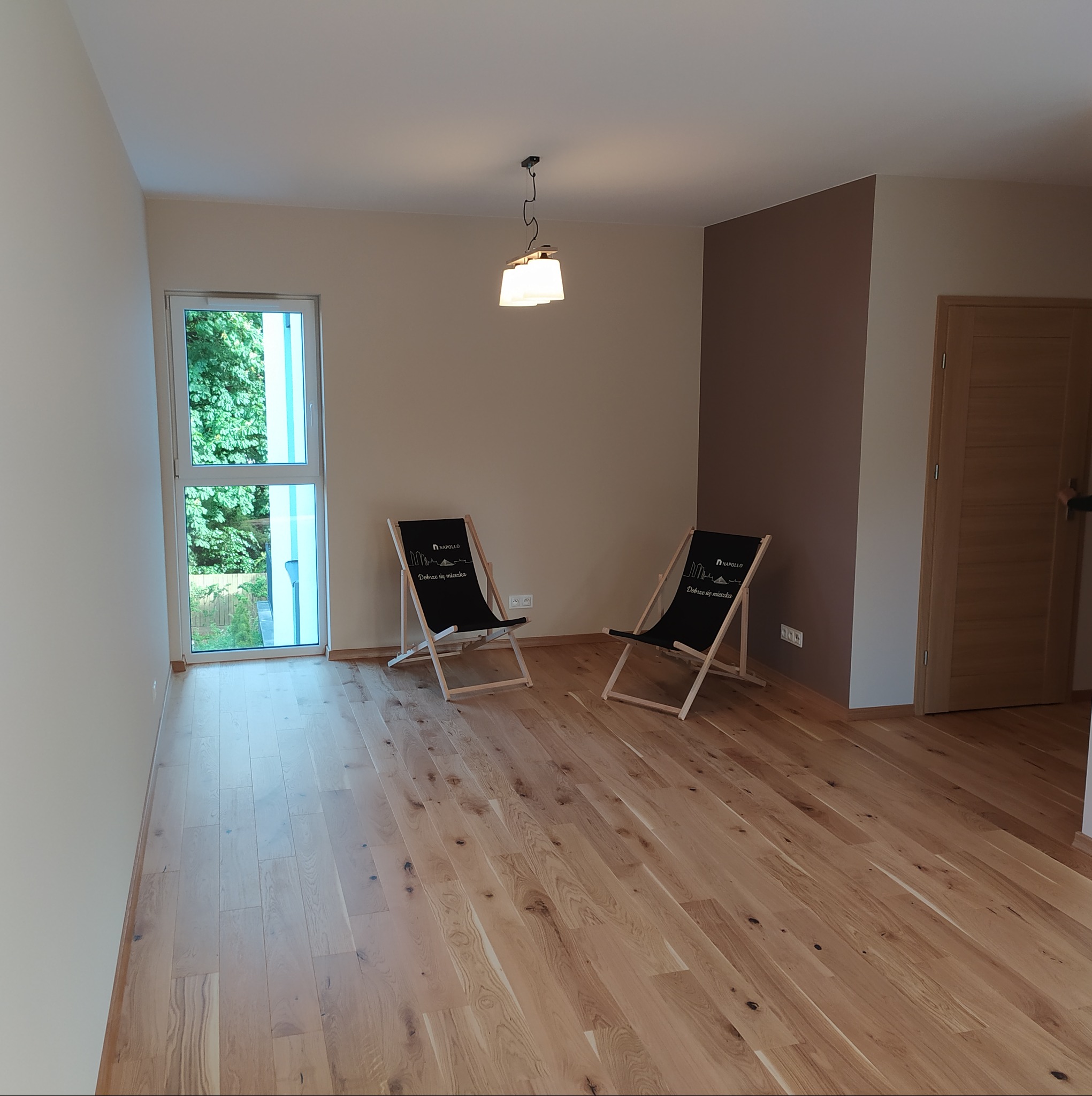 Jasne, drewniane panele podłogowe w pustym pokoju z oknem i dwoma leżakami z napisem 'Dobrze się mieszka'. Ściany w odcieniach beżu i brązu.