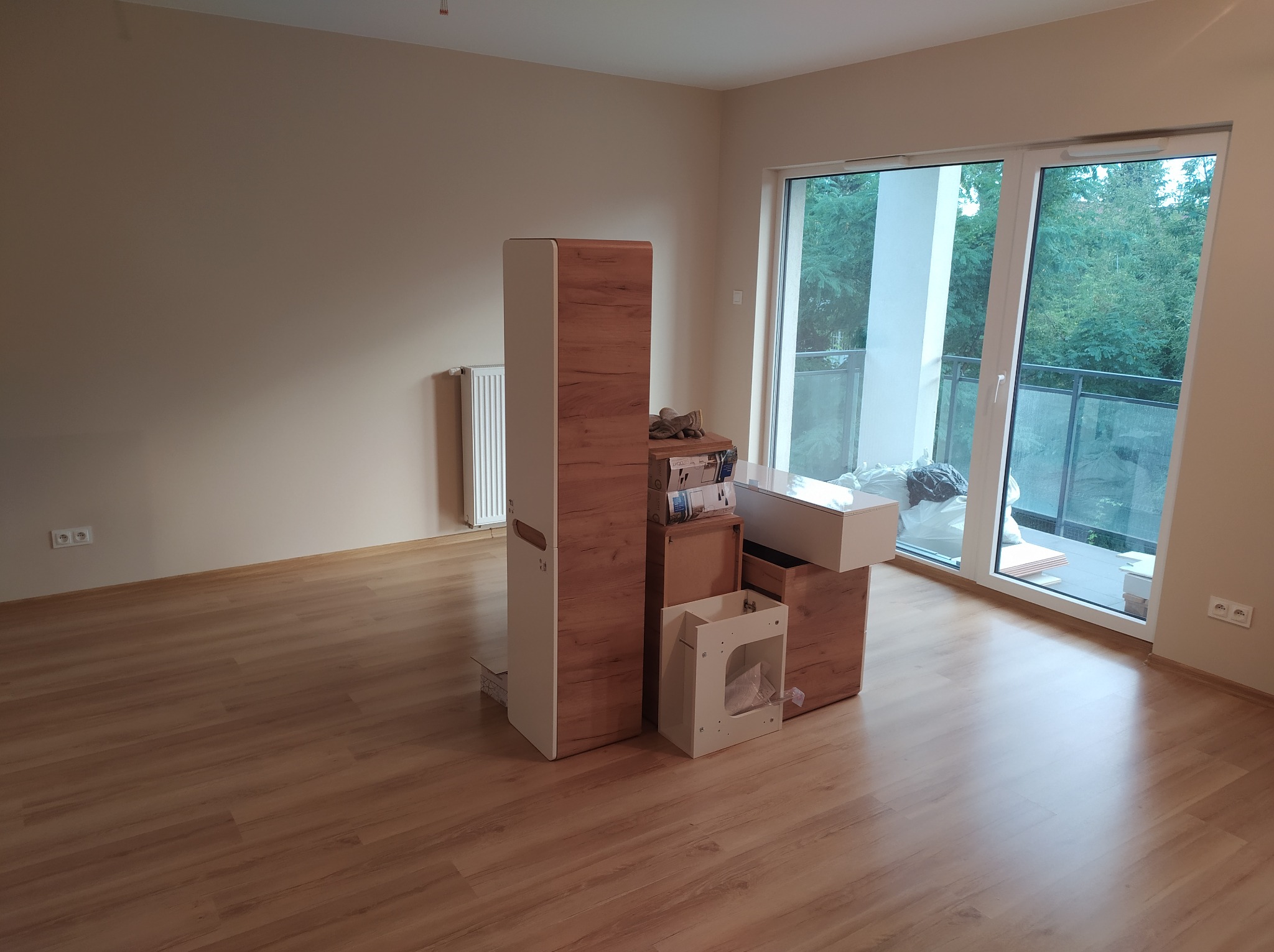 Puste, jasne wnętrze pokoju z parkietem i beżowymi ścianami po remoncie. Widoczne okno balkonowe i meble do złożenia. Ściany gładkie, gotowe do dekoracji.