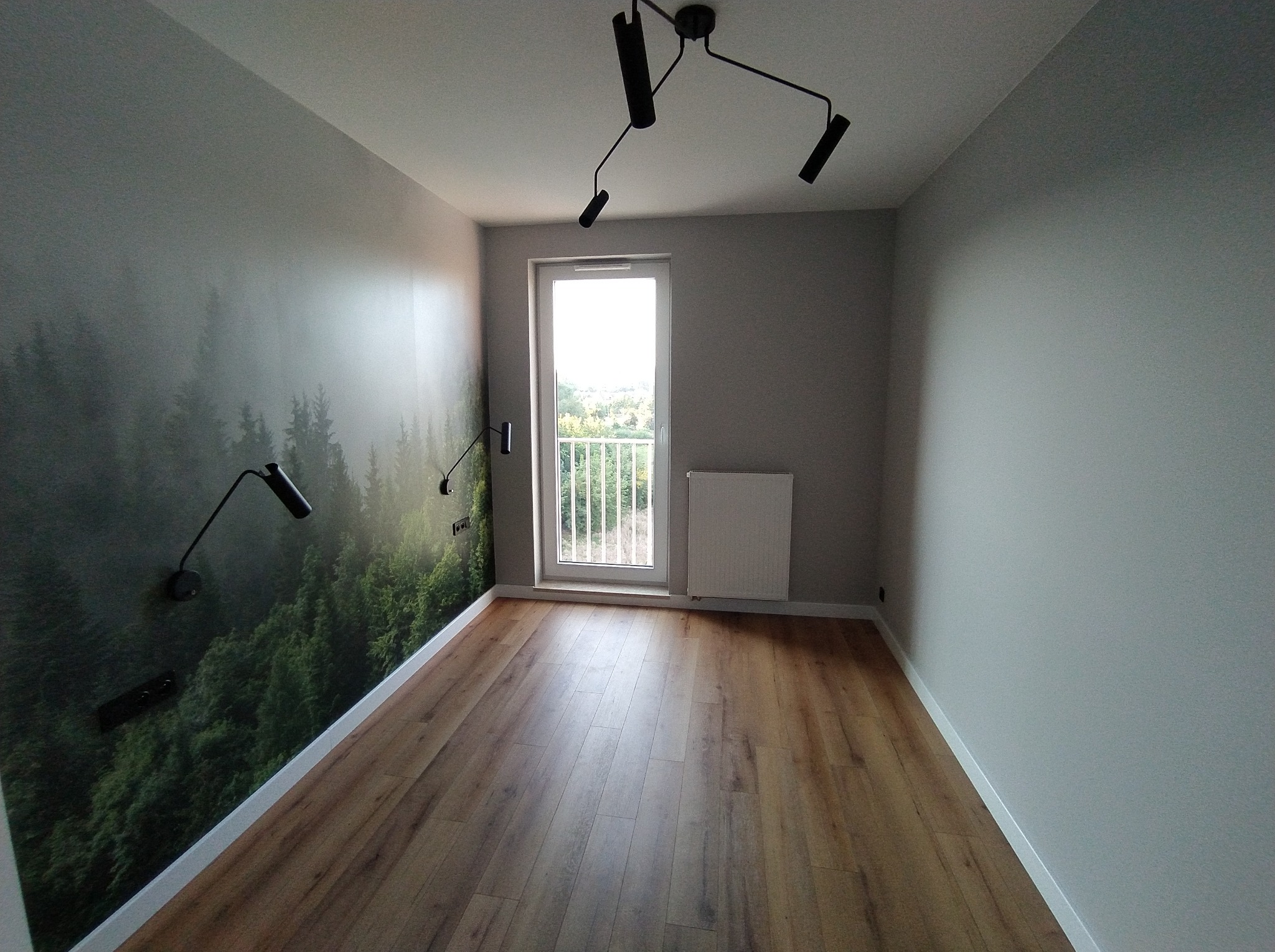 Wnętrze pokoju po remoncie: gładkie, szare ściany, fototapeta z motywem lasu, drewniana podłoga i nowoczesna lampa sufitowa.