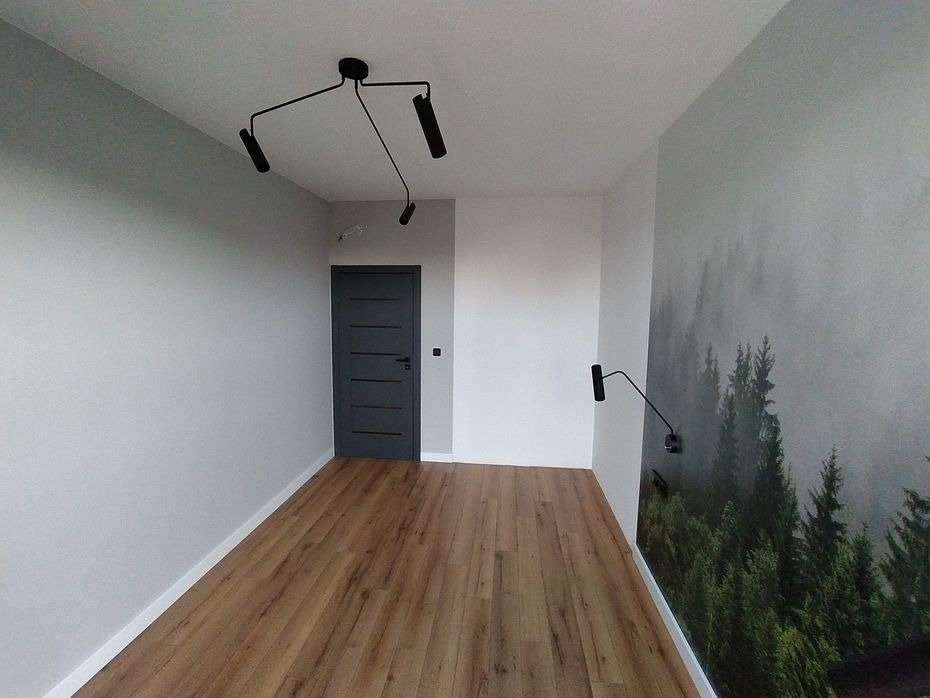 Wnętrze pokoju po remoncie: ściany pomalowane na szaro, z drewnianą podłogą i fototapetą z motywem lasu. Nowoczesne oświetlenie sufitowe i ścienne.