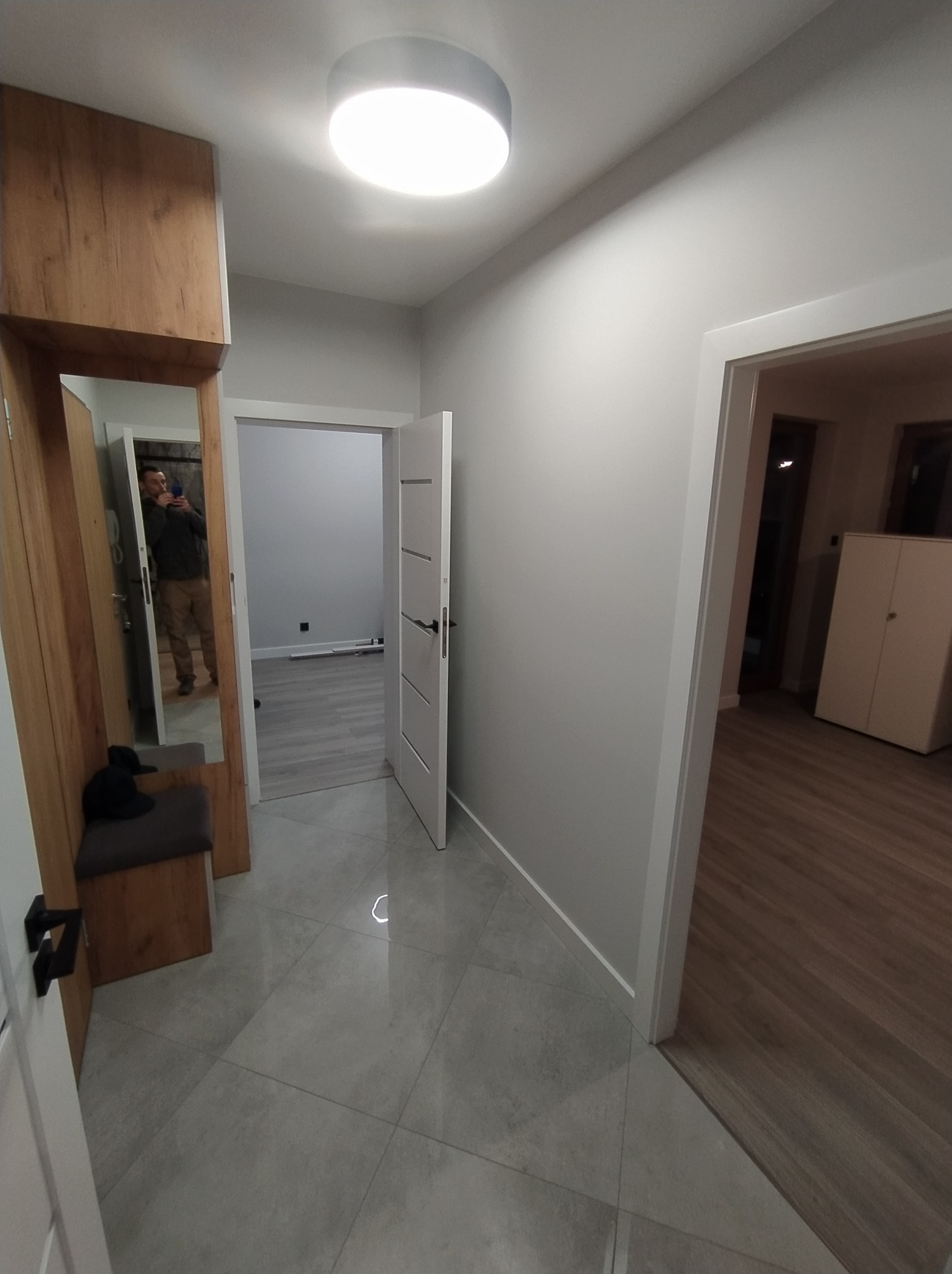 Jasne wnętrze po remoncie: gładkie, szare ściany, białe drzwi, błyszcząca podłoga z płytek. W lustrze odbicie osoby robiącej zdjęcie.