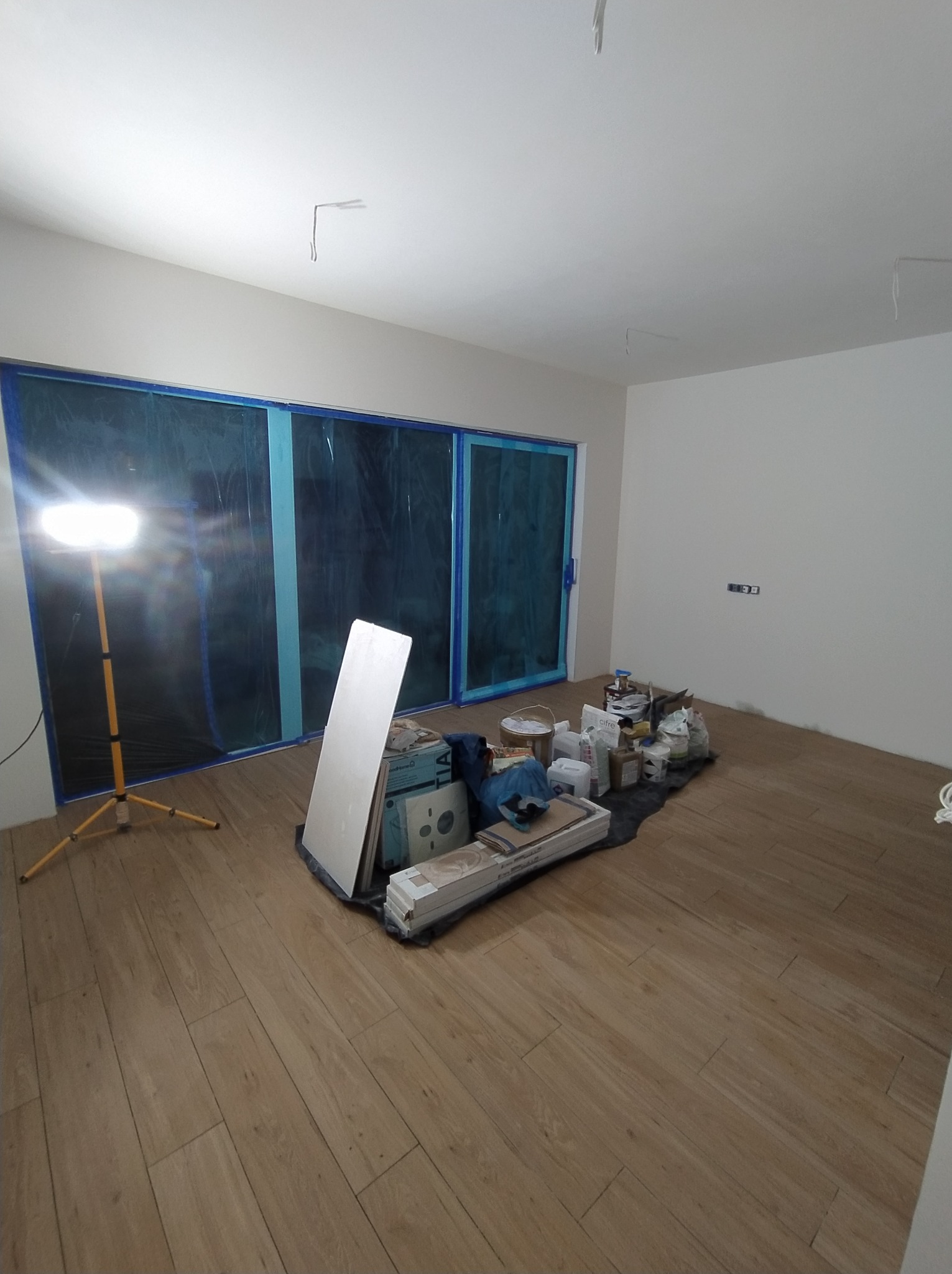 Pokój w trakcie prac wykończeniowych: zabezpieczone okna folią, podłoga wyłożona panelami, widoczny reflektor i narzędzia przygotowane do malowania ścian.