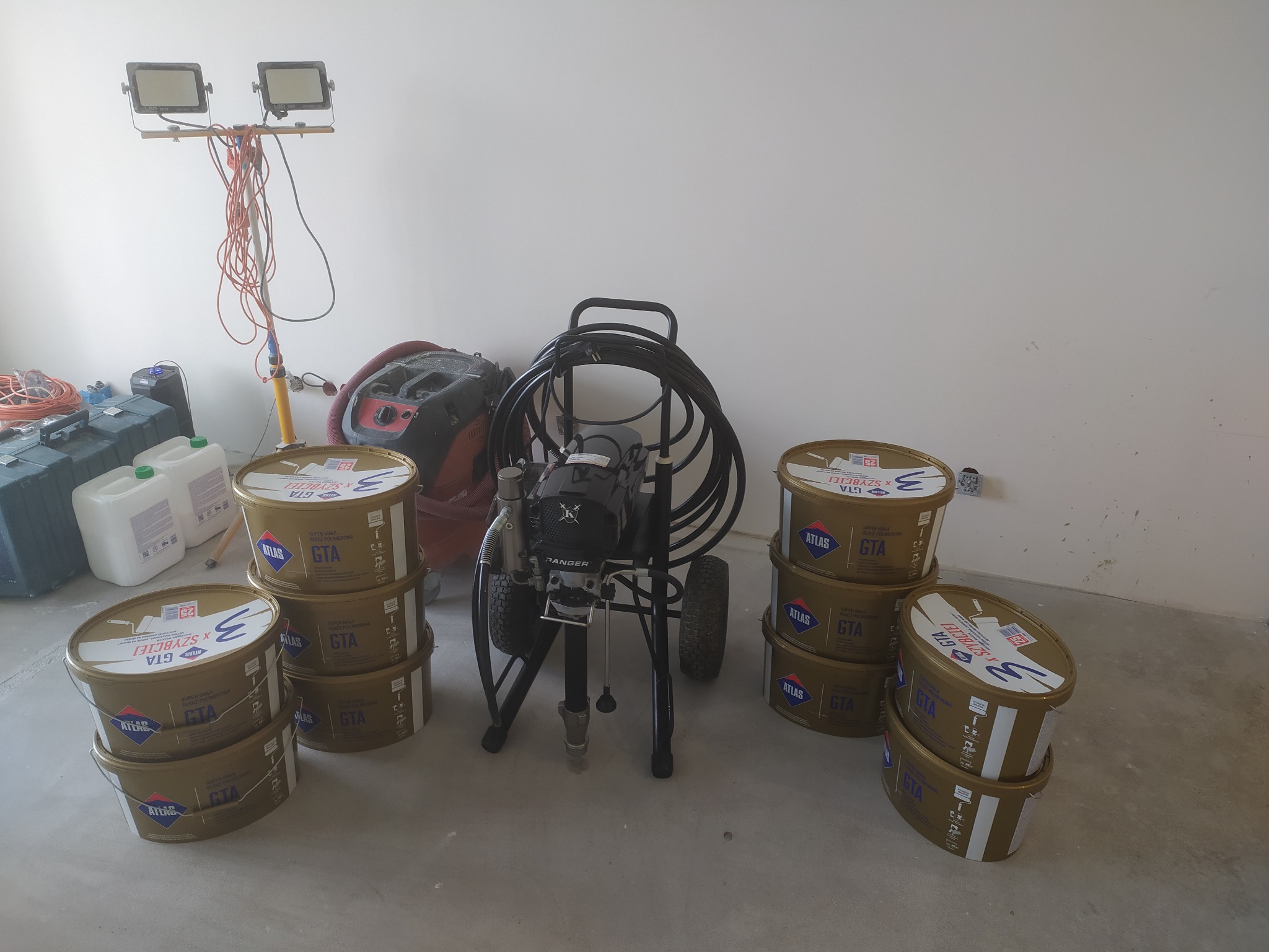 Agregat malarski stoi obok wiader z masą wykończeniową, w tle lampa budowlana i odkurzacz. Sprzęt gotowy do aplikacji w jasnym, pustym pomieszczeniu.