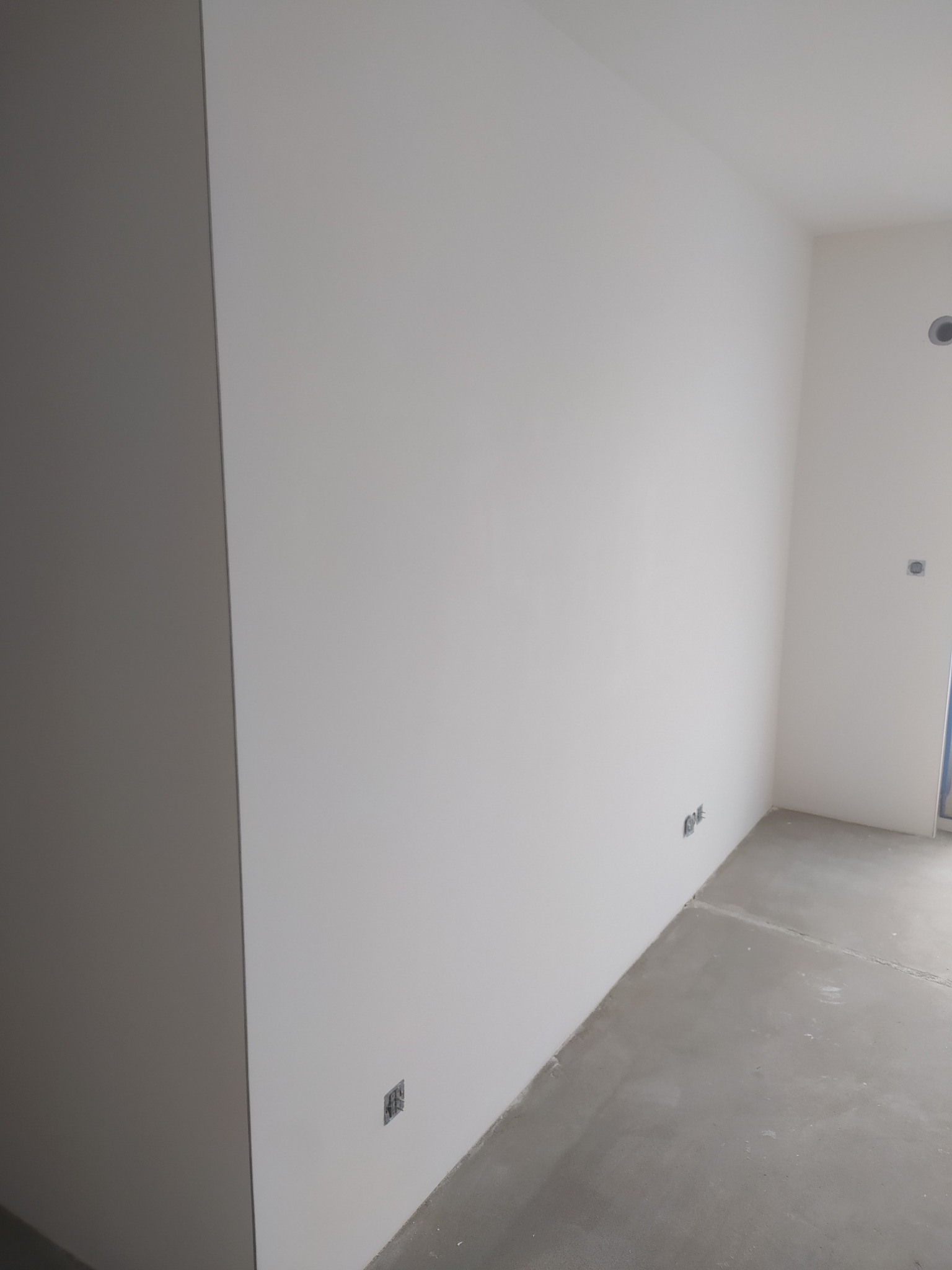 Biała, gładka ściana po szpachlowaniu, z widocznymi gniazdkami elektrycznymi i betonową podłogą. Minimalistyczny kadr, neutralne oświetlenie.