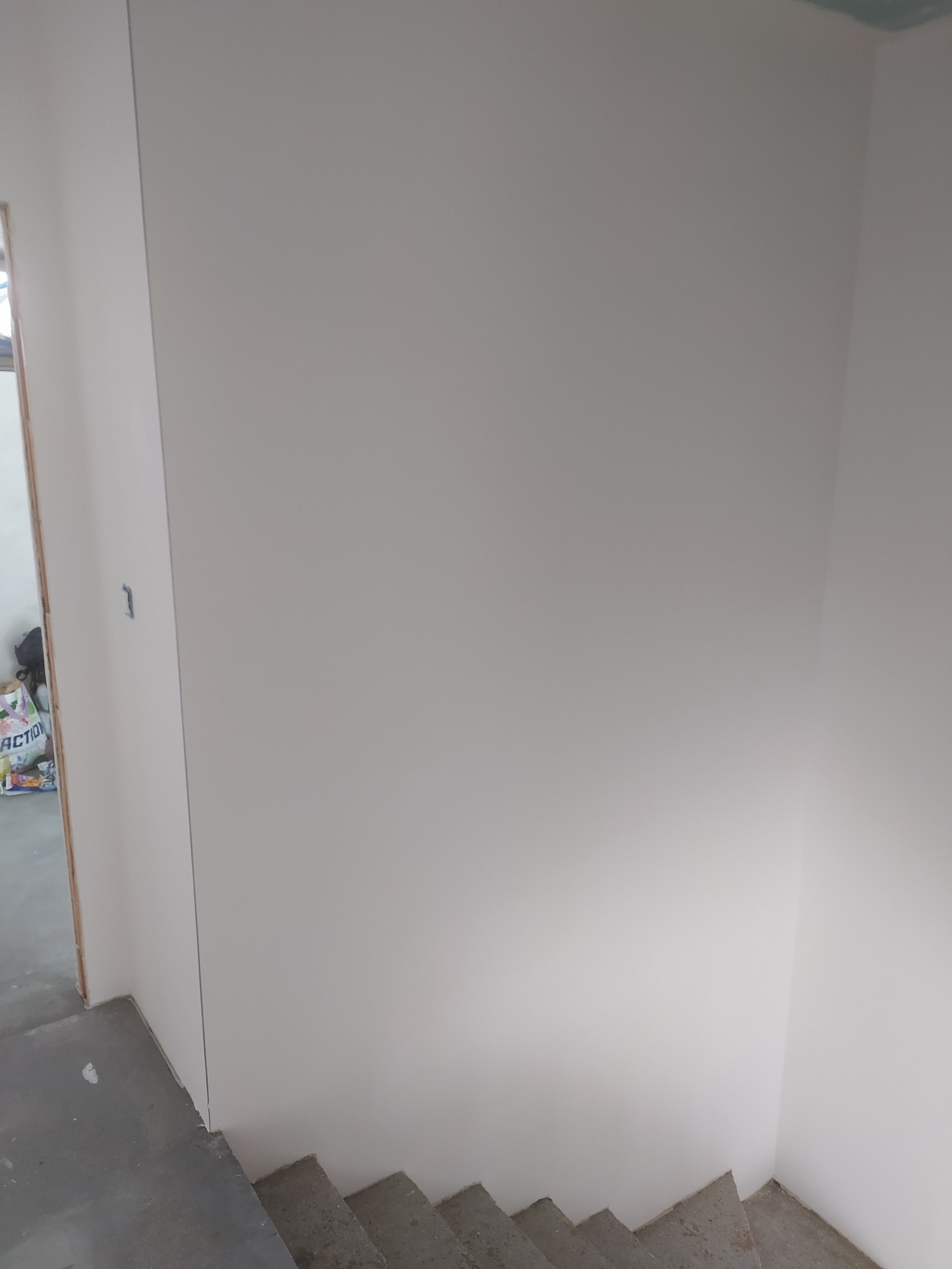 Ściana po nałożeniu jasnej, gładkiej warstwy wykończeniowej, widoczne schody w surowym stanie i fragment torby w tle.