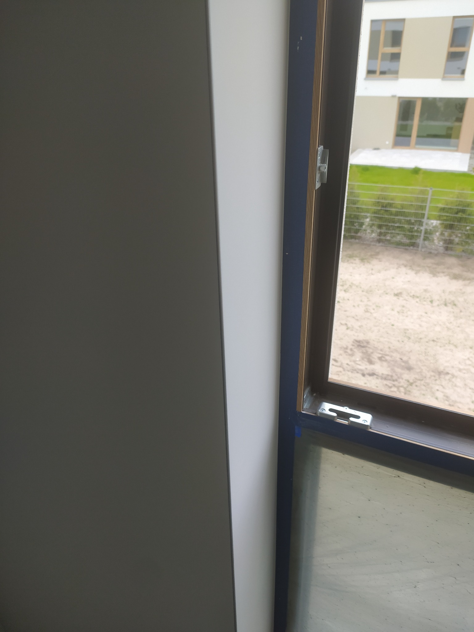 Fragment wykończonego wnętrza: gładka, biała ściana sąsiaduje z oknem z niebieską ramą, widok na zewnątrz z zielenią i budynkiem w tle.