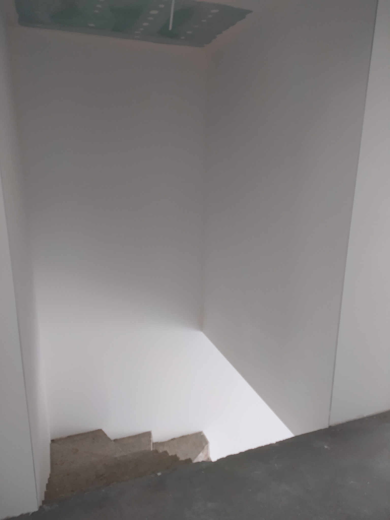 Wnętrze w trakcie wykańczania: białe ściany, surowy strop z widocznymi instalacjami i betonowe schody. Widok z góry na klatkę schodową.