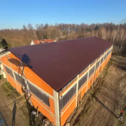 Widok z góry na nowo wybudowaną halę z brązowym dachem z blachy trapezowej, pomarańczowe ściany z pustaków, w tle drzewa bez liści i błękitne niebo, teren wokół hali częściowo zagospodarowany.