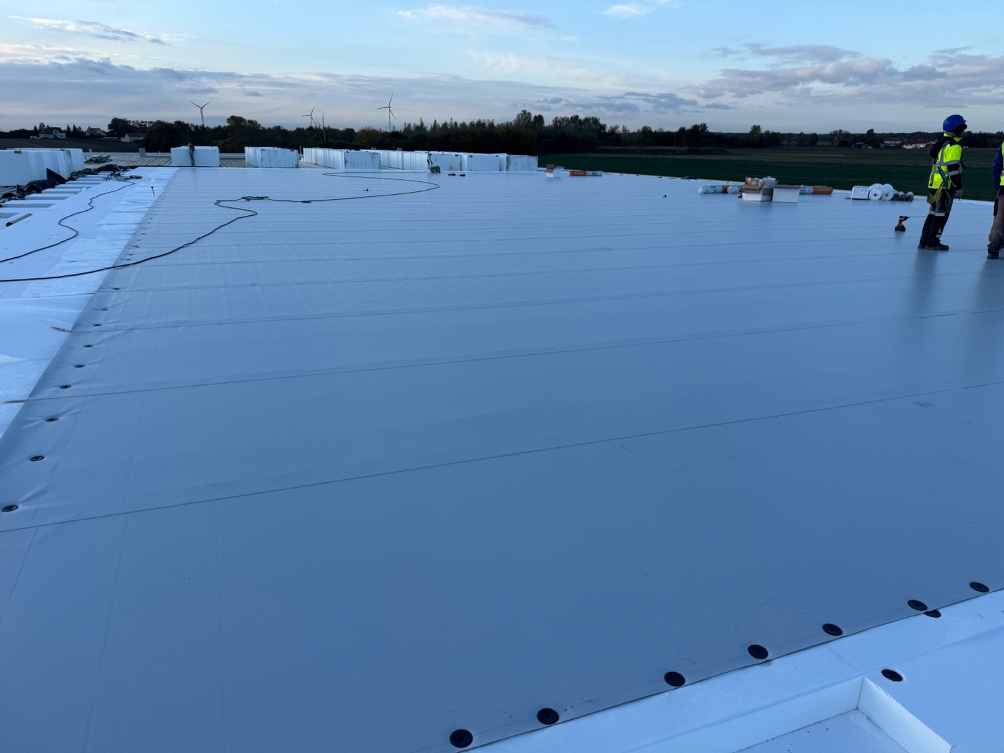 Rozległy, jasnoszary dach hali przemysłowej w trakcie montażu, widoczne czarne okrągłe mocowania, częściowo ułożona izolacja i dwóch pracowników w odblaskowych kamizelkach, w tle turbiny wiatrowe...