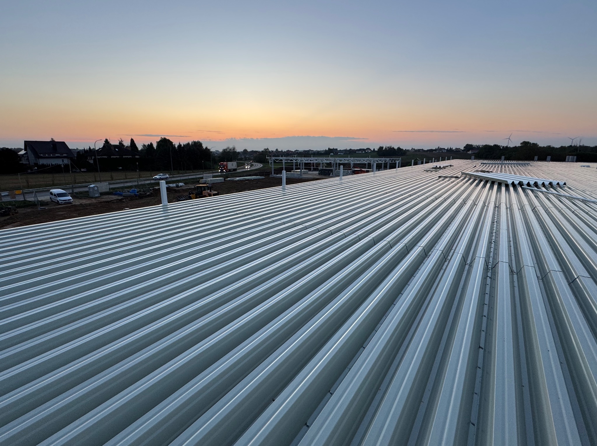 Widok z góry na nowo wybudowaną halę z szarej blachy trapezowej na tle zachodzącego słońca, widoczne elementy konstrukcji stalowej oraz droga i zabudowania w oddali.