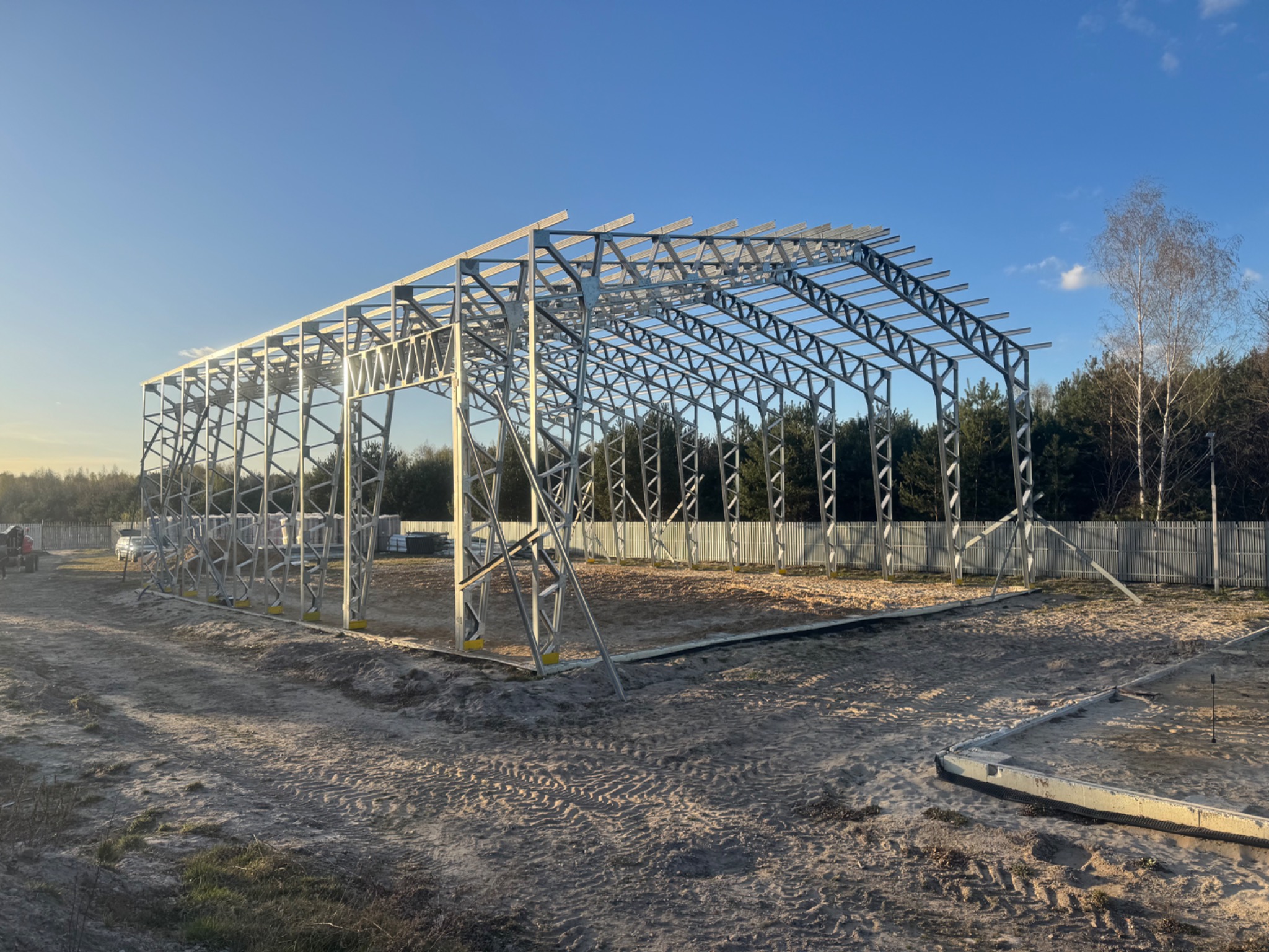 Stalowa konstrukcja szkieletowa hali w trakcie budowy, widok z zewnątrz na tle lasu i błękitnego nieba, fundamenty częściowo wylane, widoczne ślady opon na piasku.