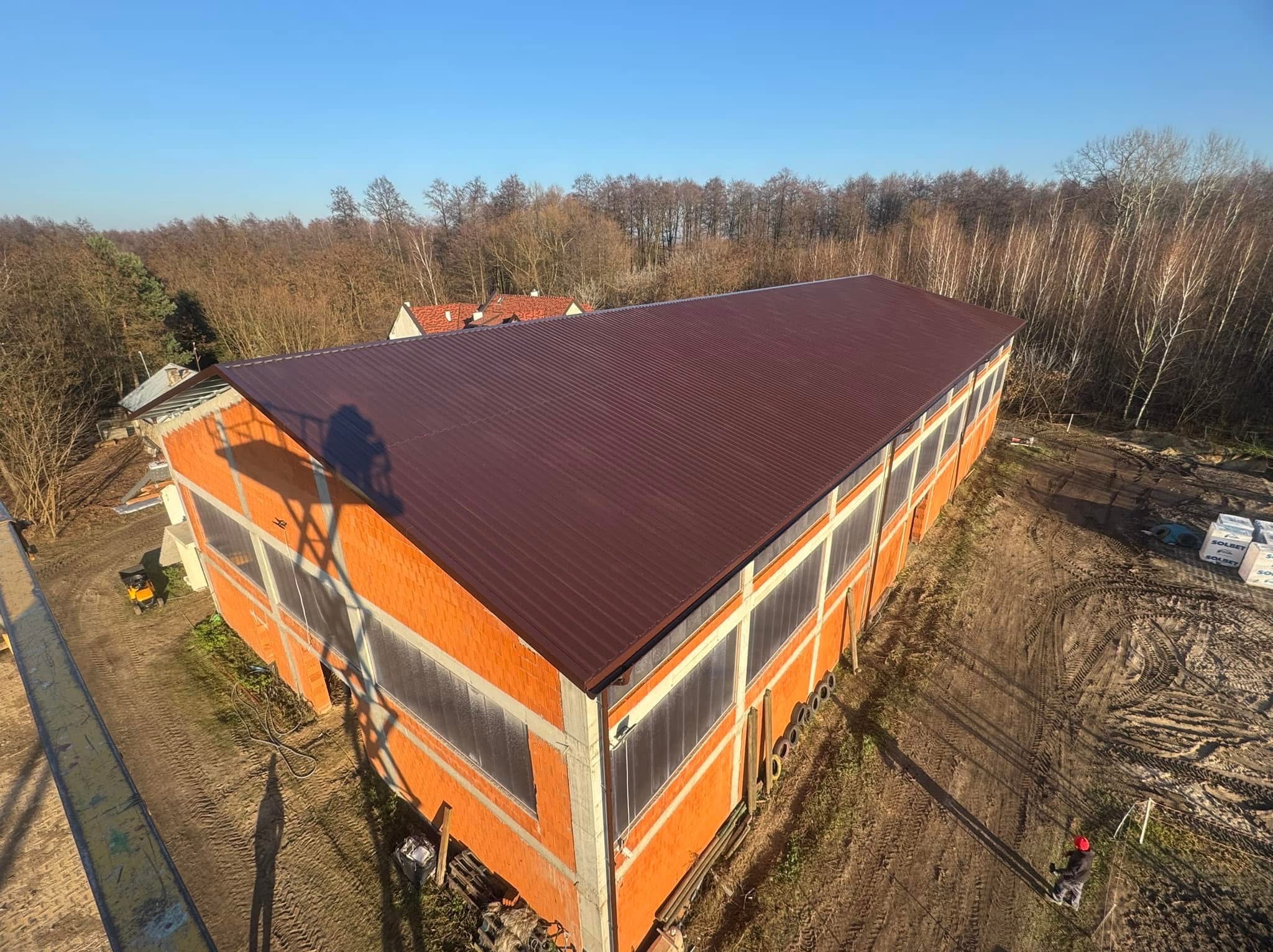Widok z góry na nowo wybudowaną halę z brązowym dachem z blachy trapezowej, pomarańczowe ściany z pustaków, w tle drzewa bez liści i błękitne niebo, teren wokół hali częściowo zagospodarowany.