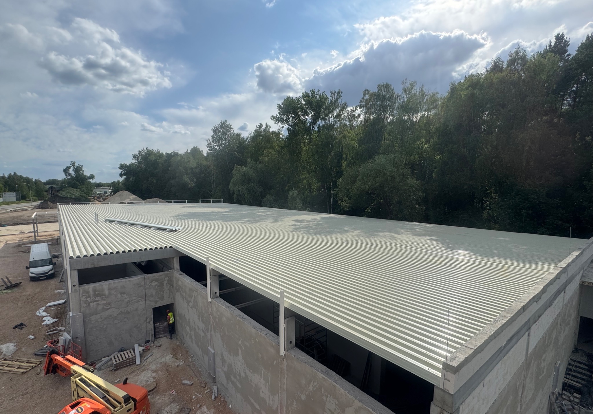 Budowa hali: widok z góry na zamontowaną, białą blachę trapezową na dachu. W tle las i błękitne niebo z chmurami. Na dole widoczny podnośnik koszowy.