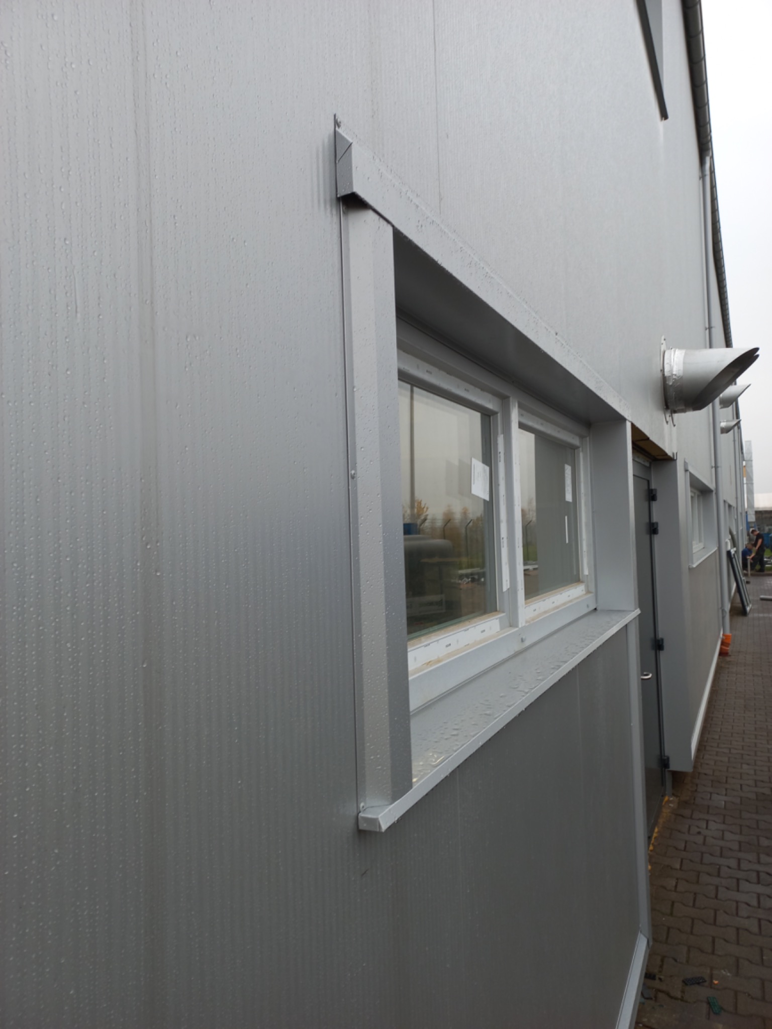 Szara hala z płyt warstwowych w deszczowy dzień, widoczny fragment okna i wentylacji.