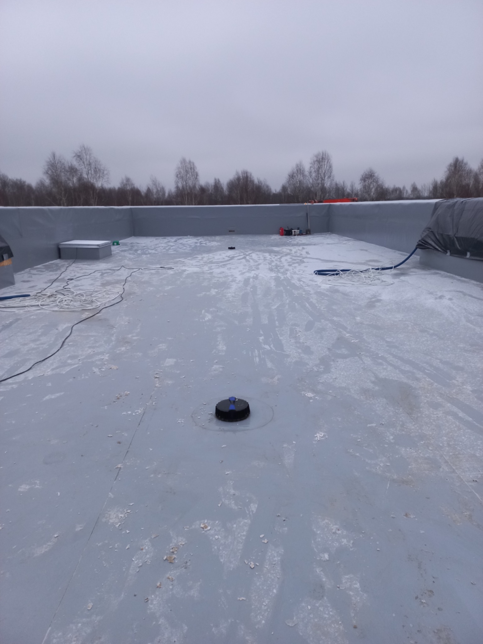 Szary, płaski dach hali przemysłowej z widocznym odpływem, częściowo pokryty białym nalotem, z elementami wyposażenia i drzewami w tle, sfotografowany w pochmurny dzień.