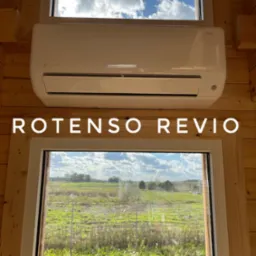 Biały klimatyzator Rotenso Revio zamontowany nad oknem w drewnianym domku, widok na zielone pole i zachmurzone niebo.