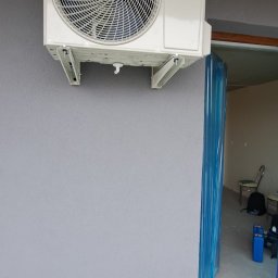 KLIMA STAR - Biały klimatyzator marki AUX zamontowany na szarej ścianie wewnątrz budynku, widoczne metalowe wsporniki i fragment okna zabezpieczonego folią ochronną.