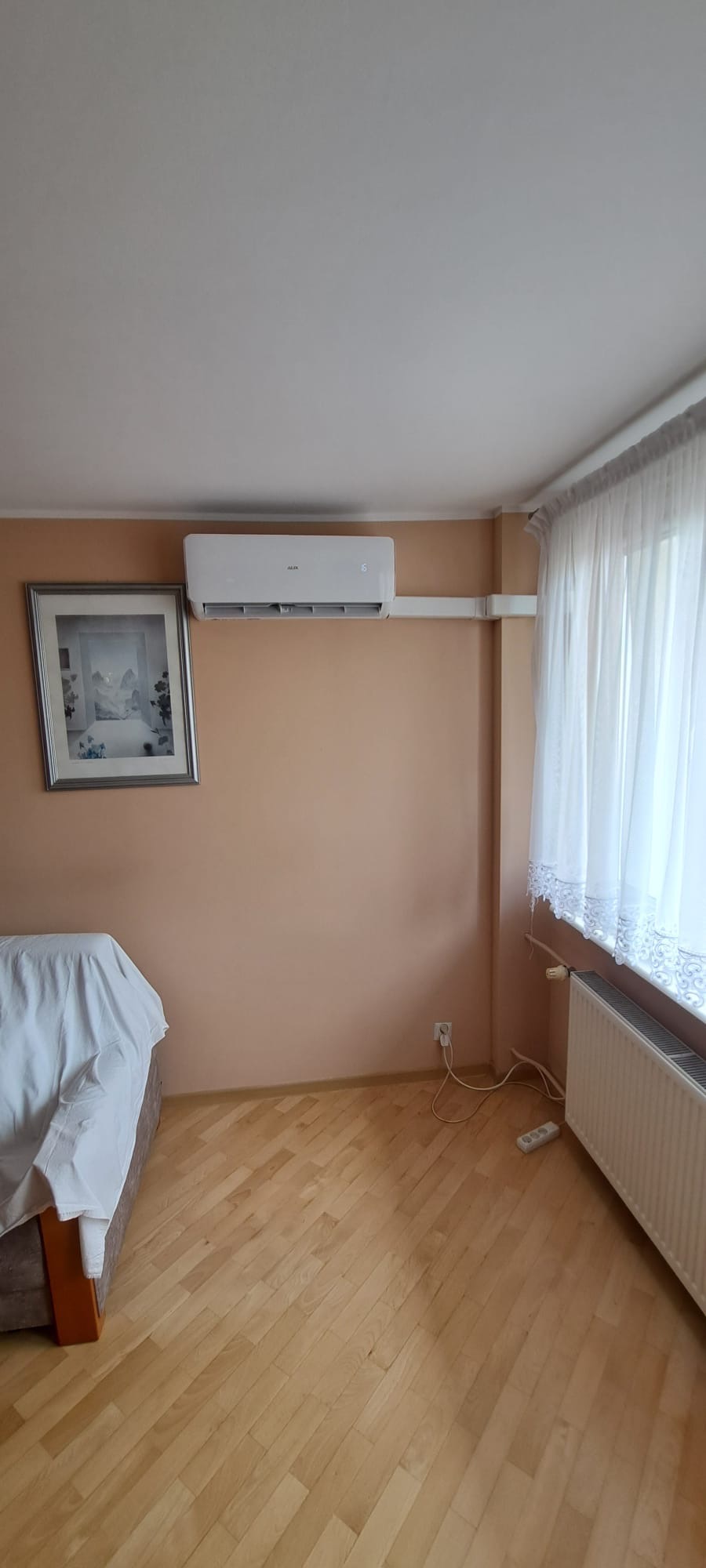 Biały klimatyzator AUX zamontowany na ścianie w pokoju o morelowych ścianach, widoczne rury instalacyjne, obraz w ramie, zasłonięte okno, drewniana podłoga.