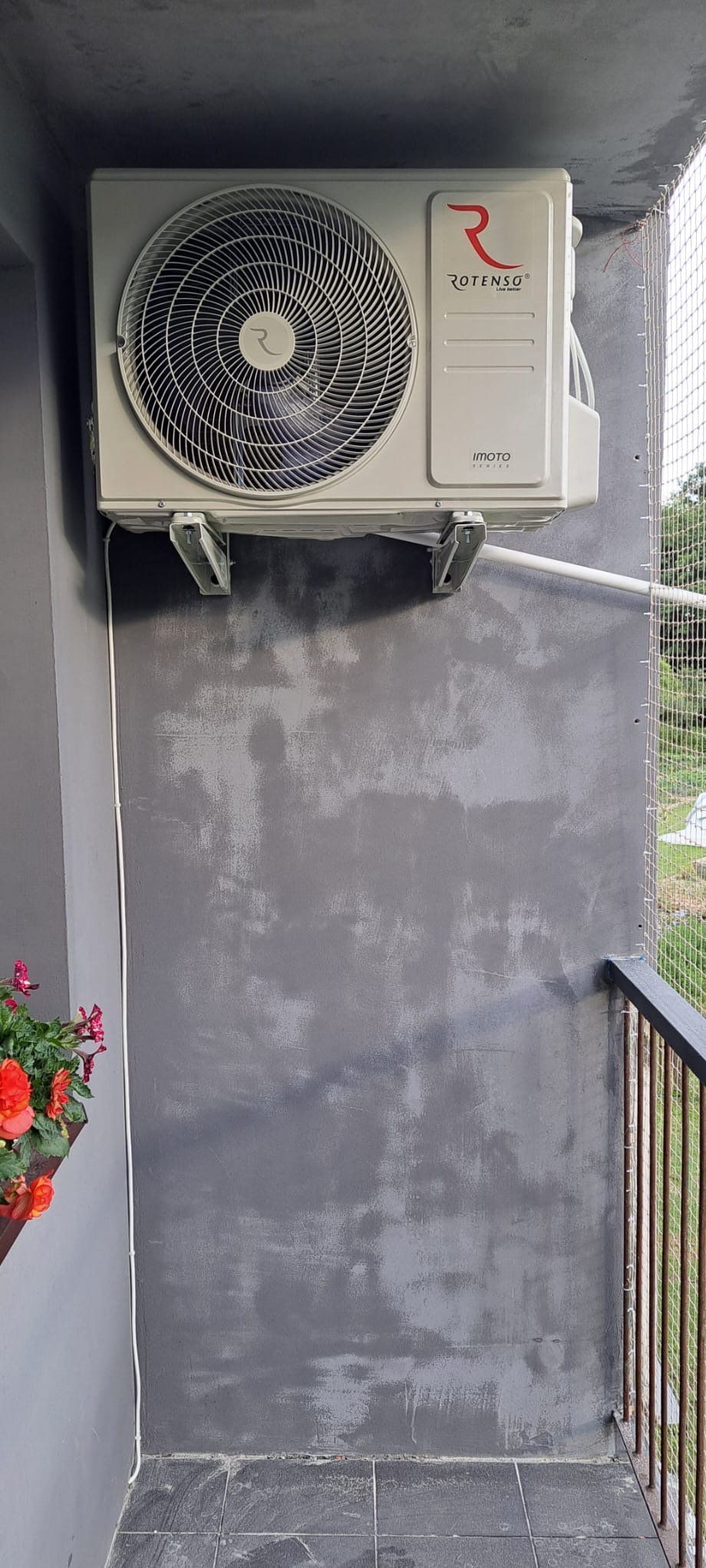 Zewnętrzna jednostka klimatyzacji Rotenso Imoto zamontowana na szarej ścianie balkonu z widoczną kratką zabezpieczającą i doniczką z czerwonymi kwiatami.