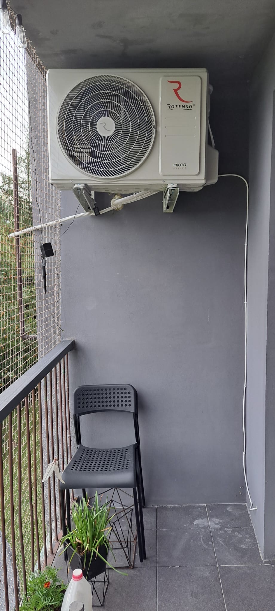 Zamontowana jednostka zewnętrzna klimatyzacji Rotenso Imoto na balkonie z szarą ścianą i czarnym krzesłem, widok z dołu.