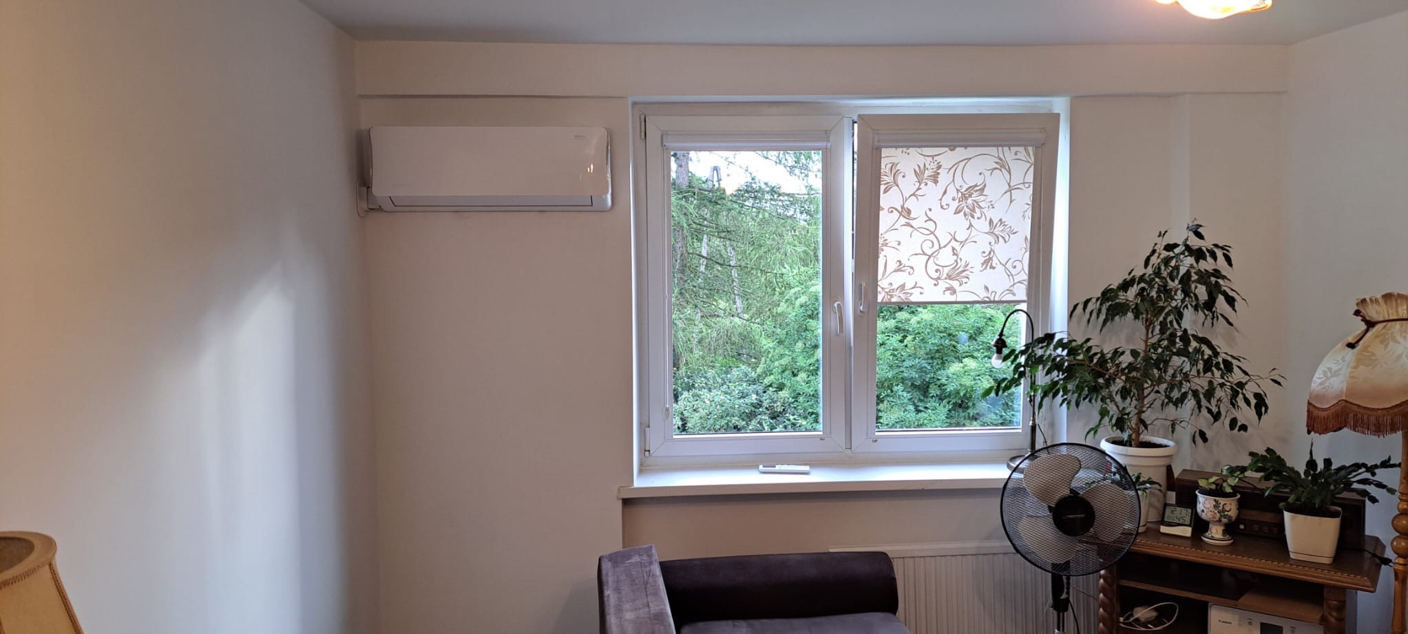 Wnętrze pokoju z zamontowanym klimatyzatorem ściennym, widok na okno z roletą w kwiaty i rośliny doniczkowe.