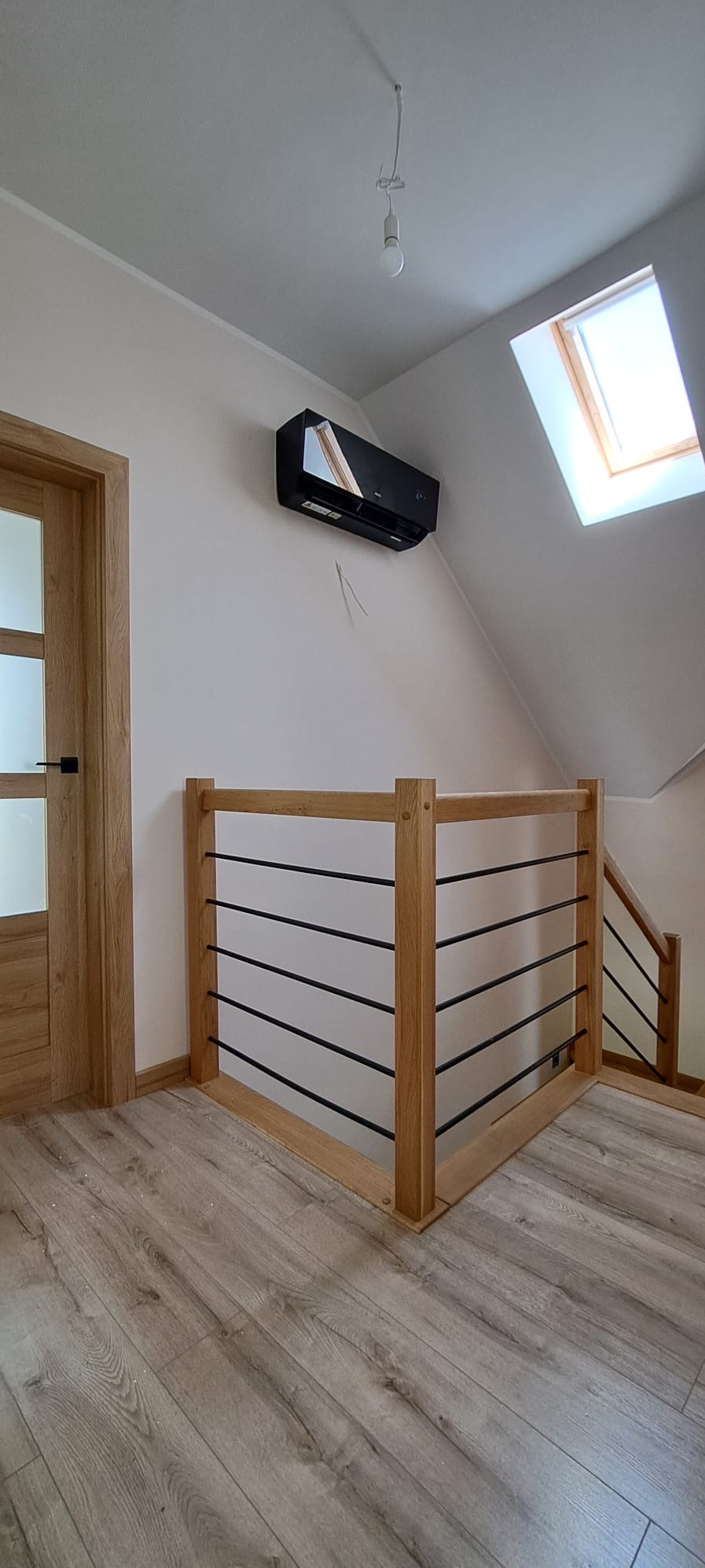 Czarna jednostka klimatyzacyjna zamontowana na ścianie w jasnym pomieszczeniu na poddaszu, widoczna drewniana balustrada schodów i okno dachowe.