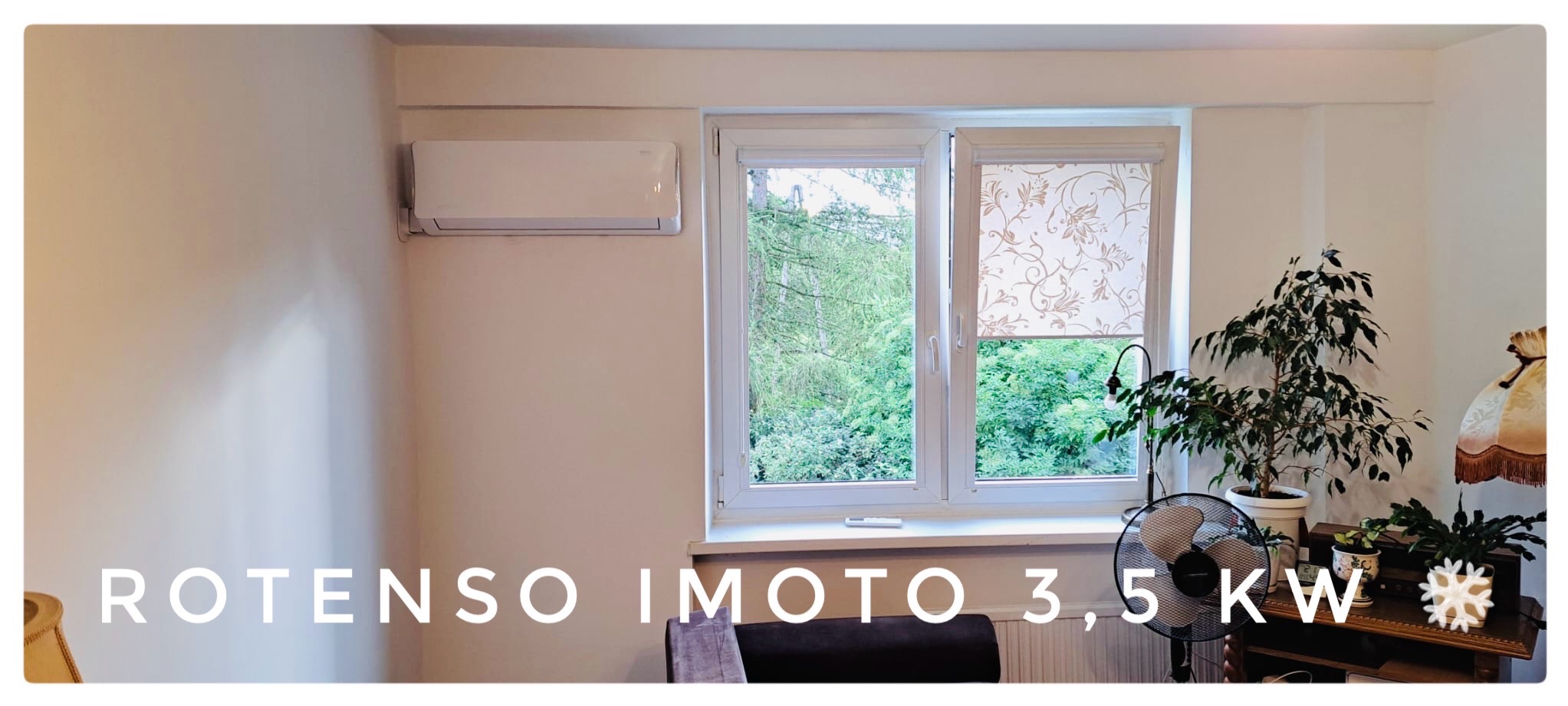 Biały klimatyzator Rotenso IMOTO 3,5 KW zamontowany na ścianie w jasnym pokoju z oknem, zasłoniętym roletą w kwiatowy wzór, widok na zieleń za oknem, dodatkowo widoczny wentylator i rośliny...
